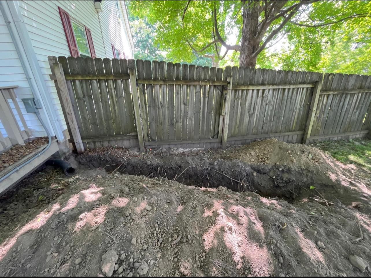 Trench dug along a wooden fence, with piles of dirt in the foreground. Building on the left, trees in background.