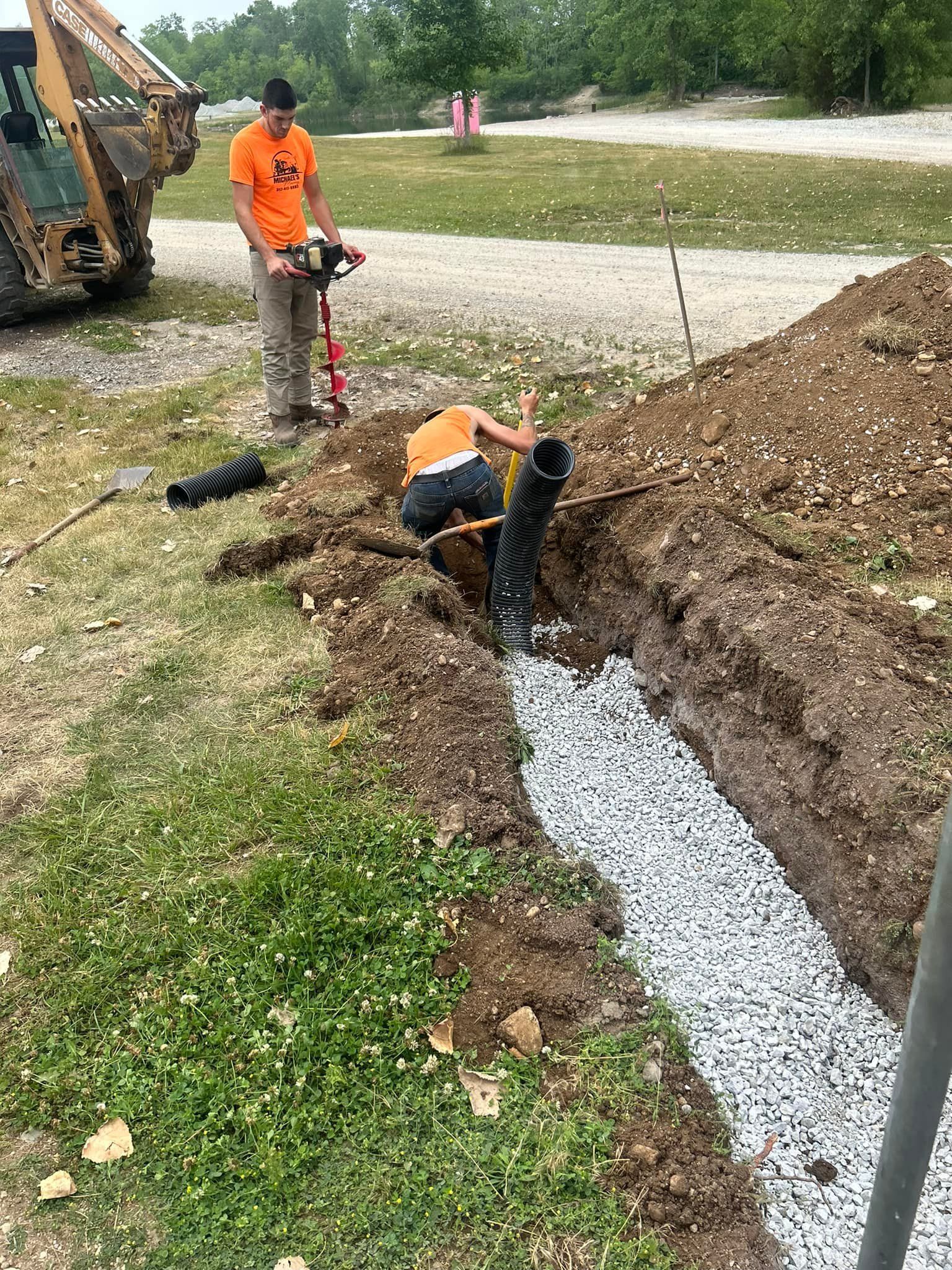 Two people installing drainage pipe in a trench filled with gravel. One uses a drill.