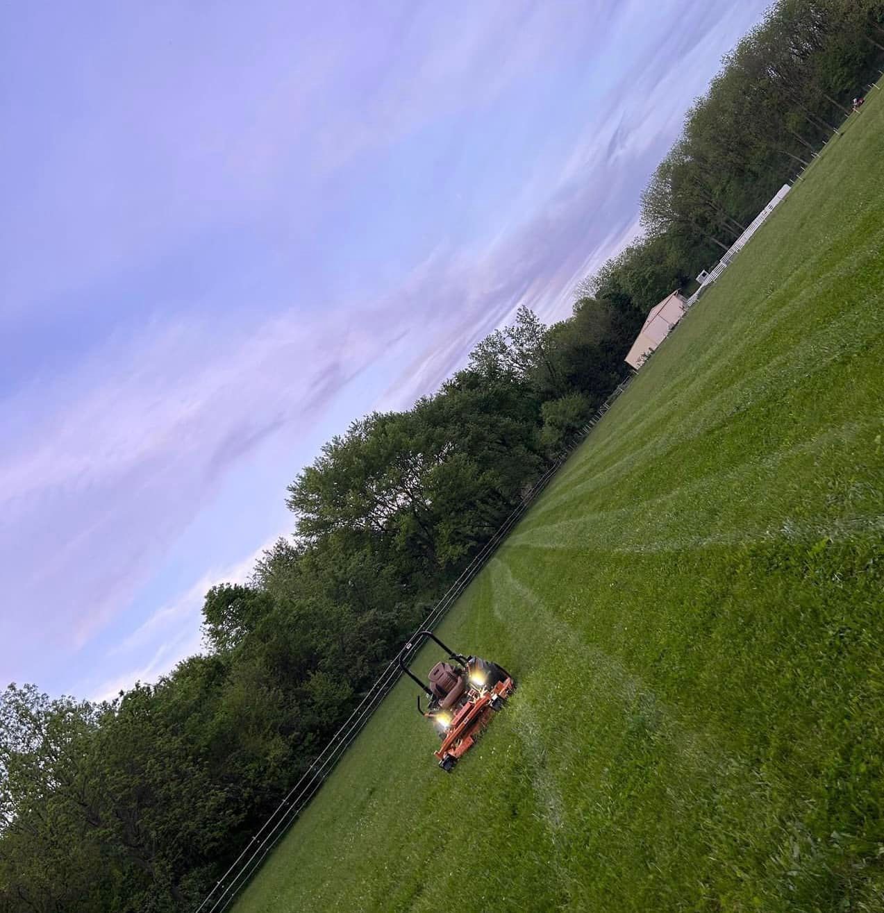 Lawn mower cutting grass in a field, with trees on one side and a cloudy sky above.