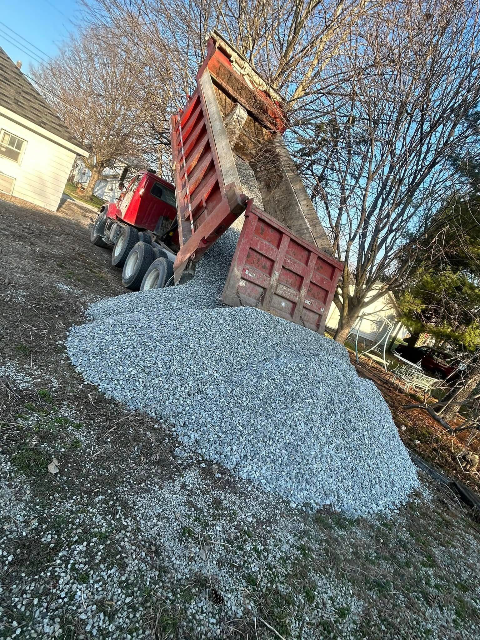 Red dump truck tilting to unload gravel onto a sloped lawn.