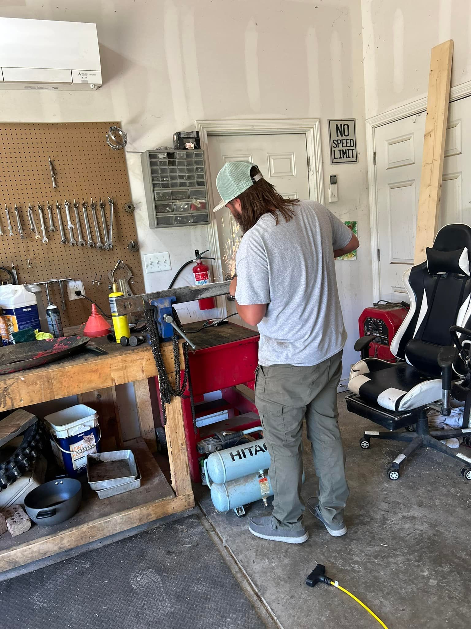 Man in a garage working on equipment. Red cart, pegboard with tools, air compressor, and workbench.