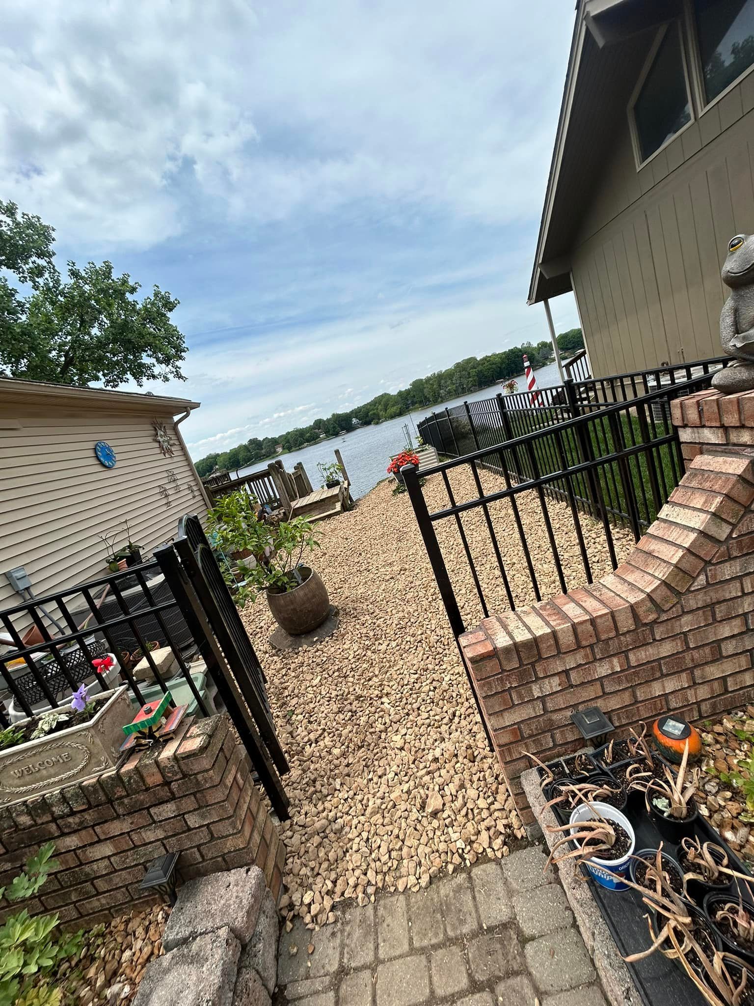 View of a lake from a brick and black iron fenced yard with a home and cloudy sky in the background.