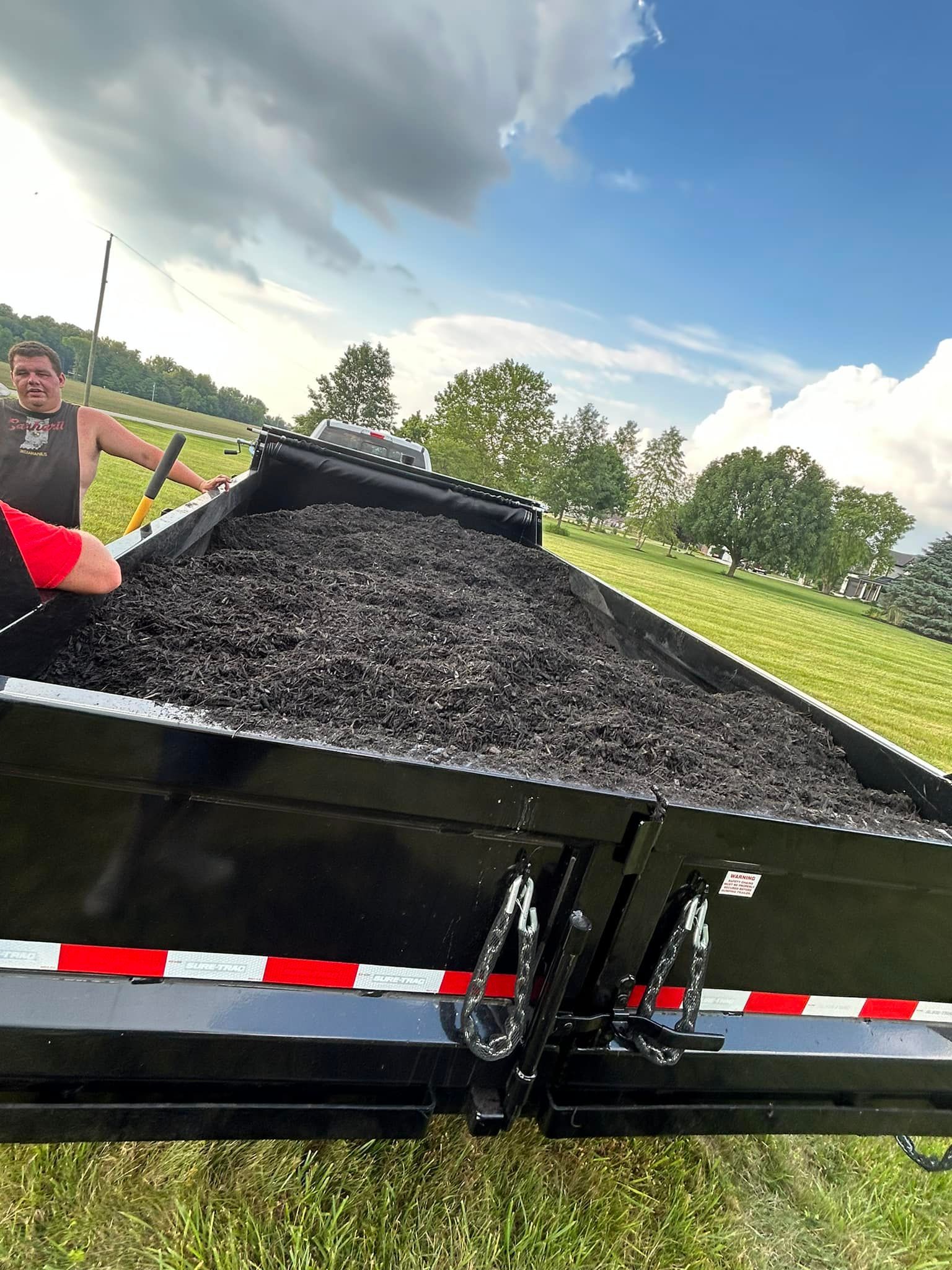 Man with shovel loading black mulch into black trailer in grassy field.