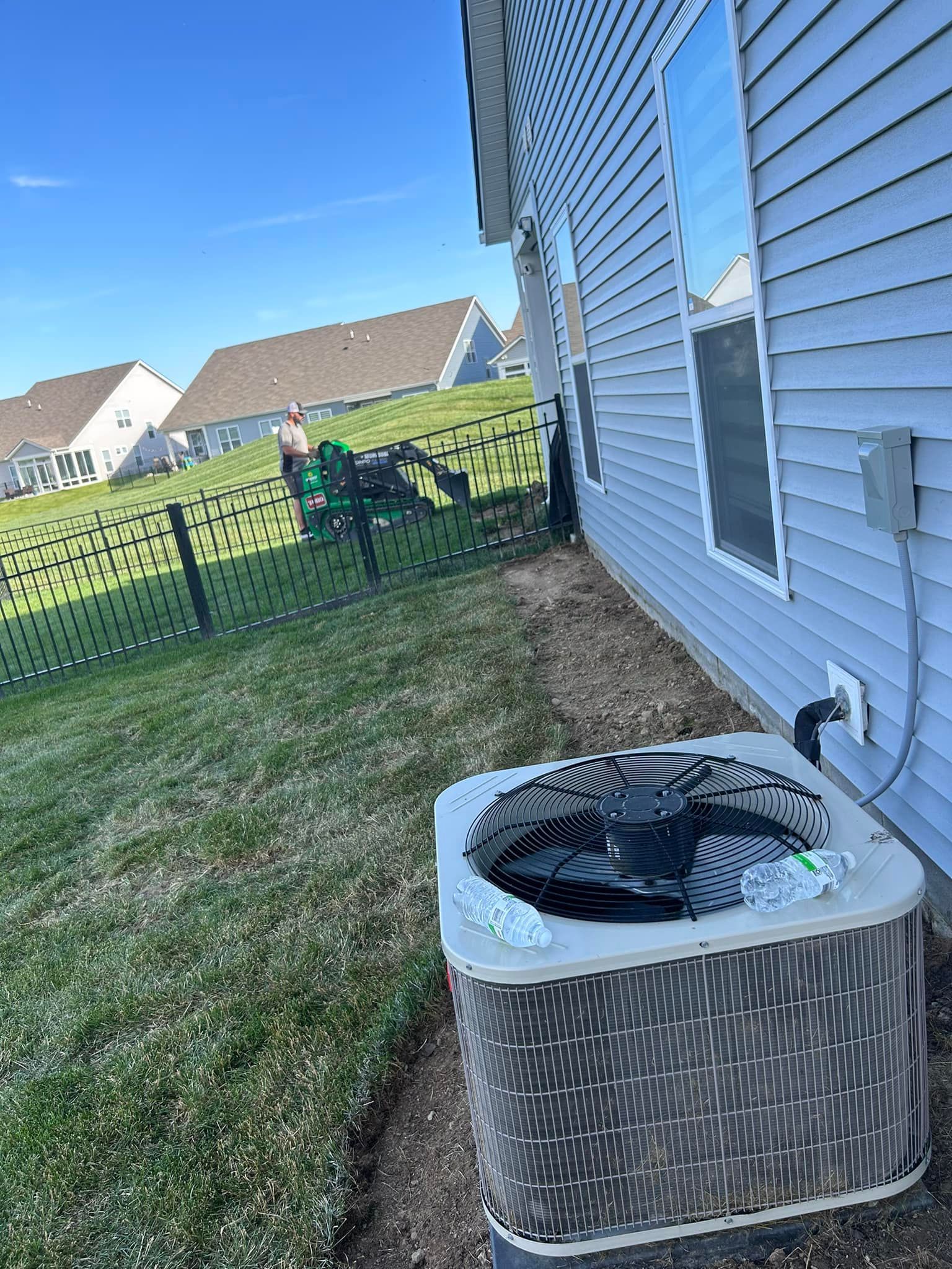 An air conditioning unit next to a house with a person in the background, blue sky.