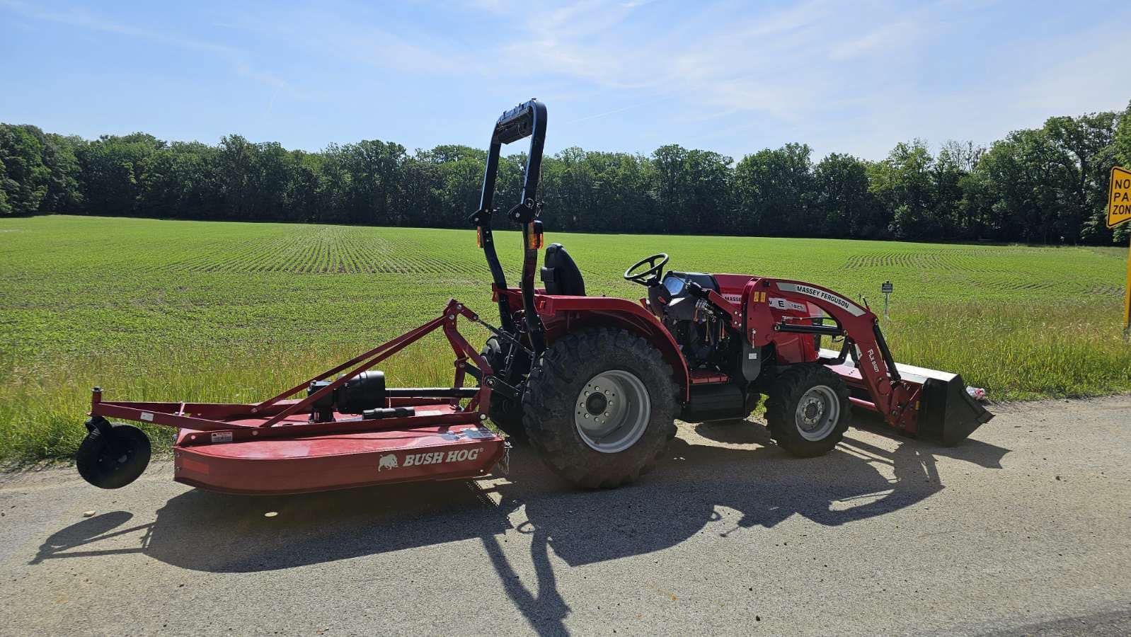 Red tractor with front loader and brush hog mower parked on a roadside, green field and trees in the background.