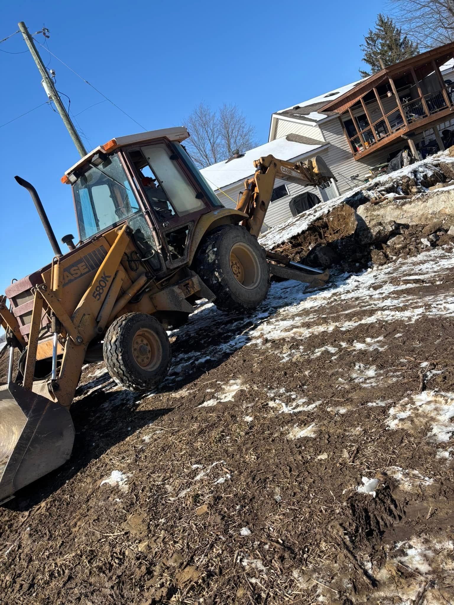 Yellow backhoe digging into muddy, snowy hillside near a house on a sunny day.