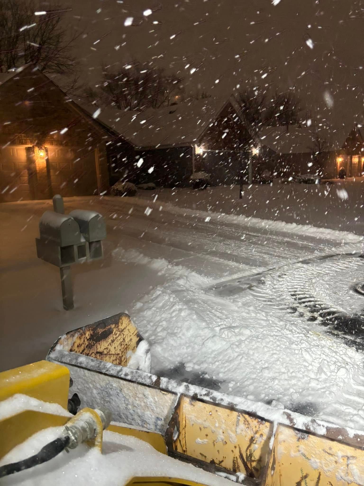 Snowplow clearing a snowy street at night, with a mailbox in the foreground and homes in the background.