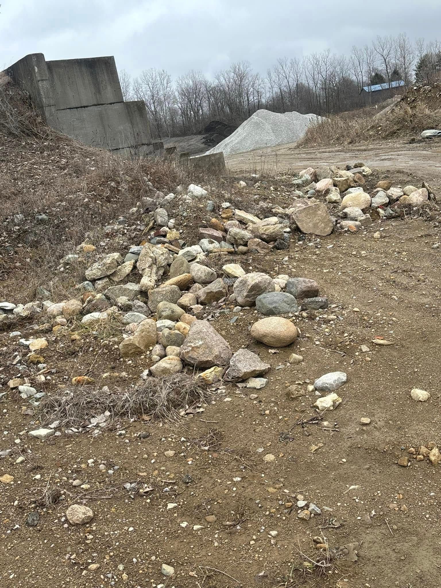 Pile of rocks in a dirt lot; concrete blocks and brush in the background under a cloudy sky.