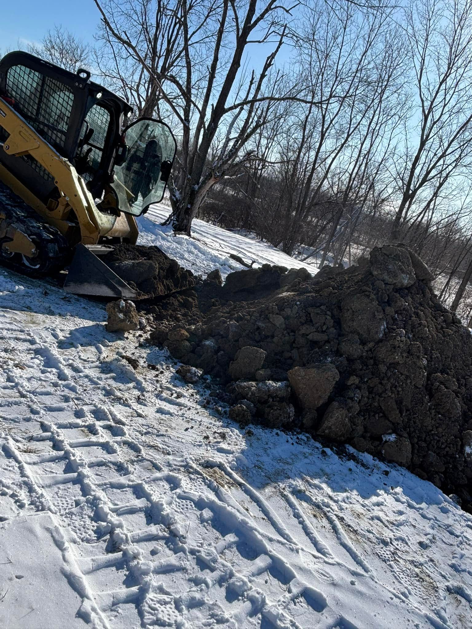 Yellow skid steer excavating dirt near trees on snowy slope.