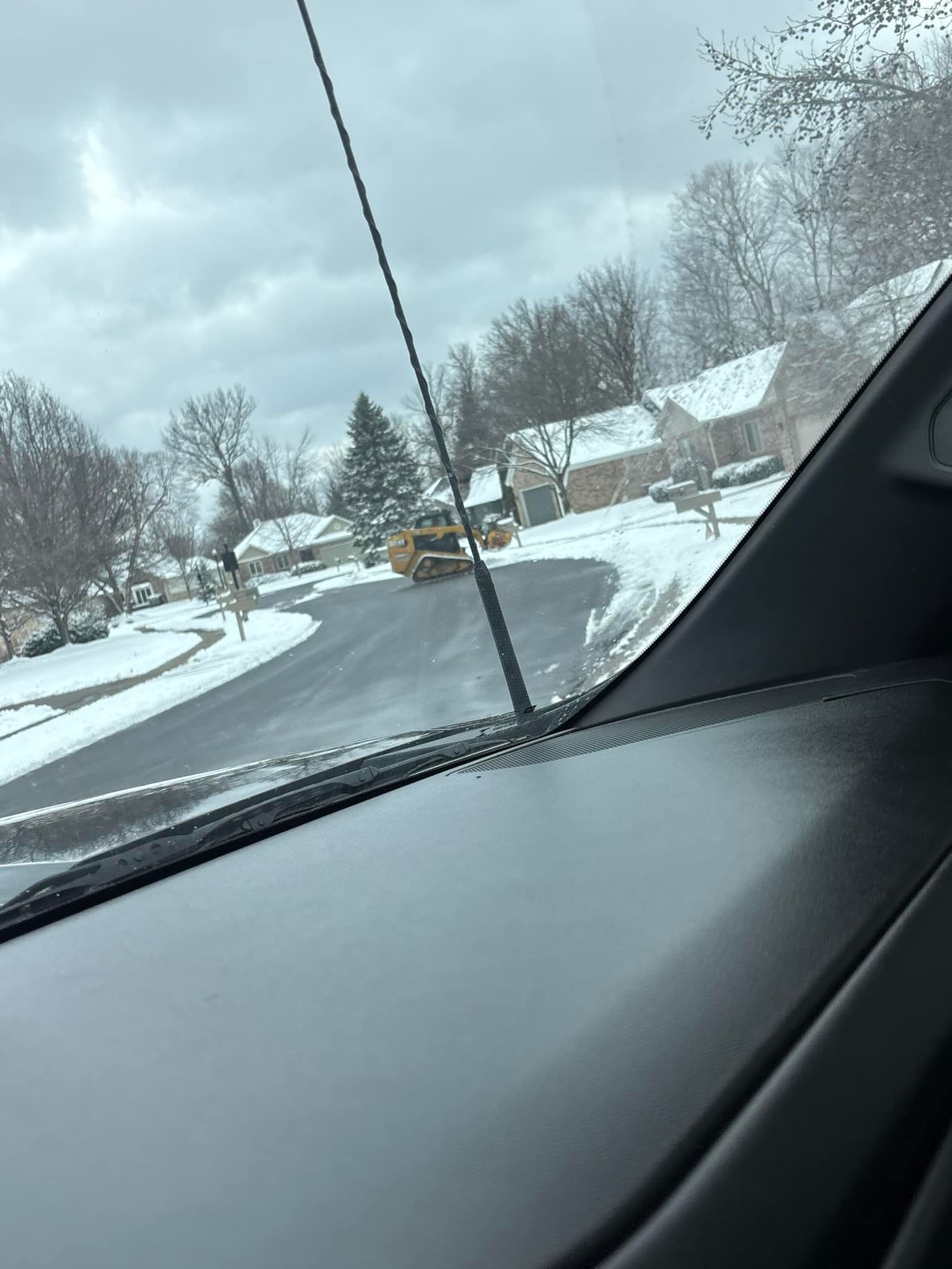 Snowy residential street seen from inside a vehicle, with a black antenna and dashboard. Overcast sky.