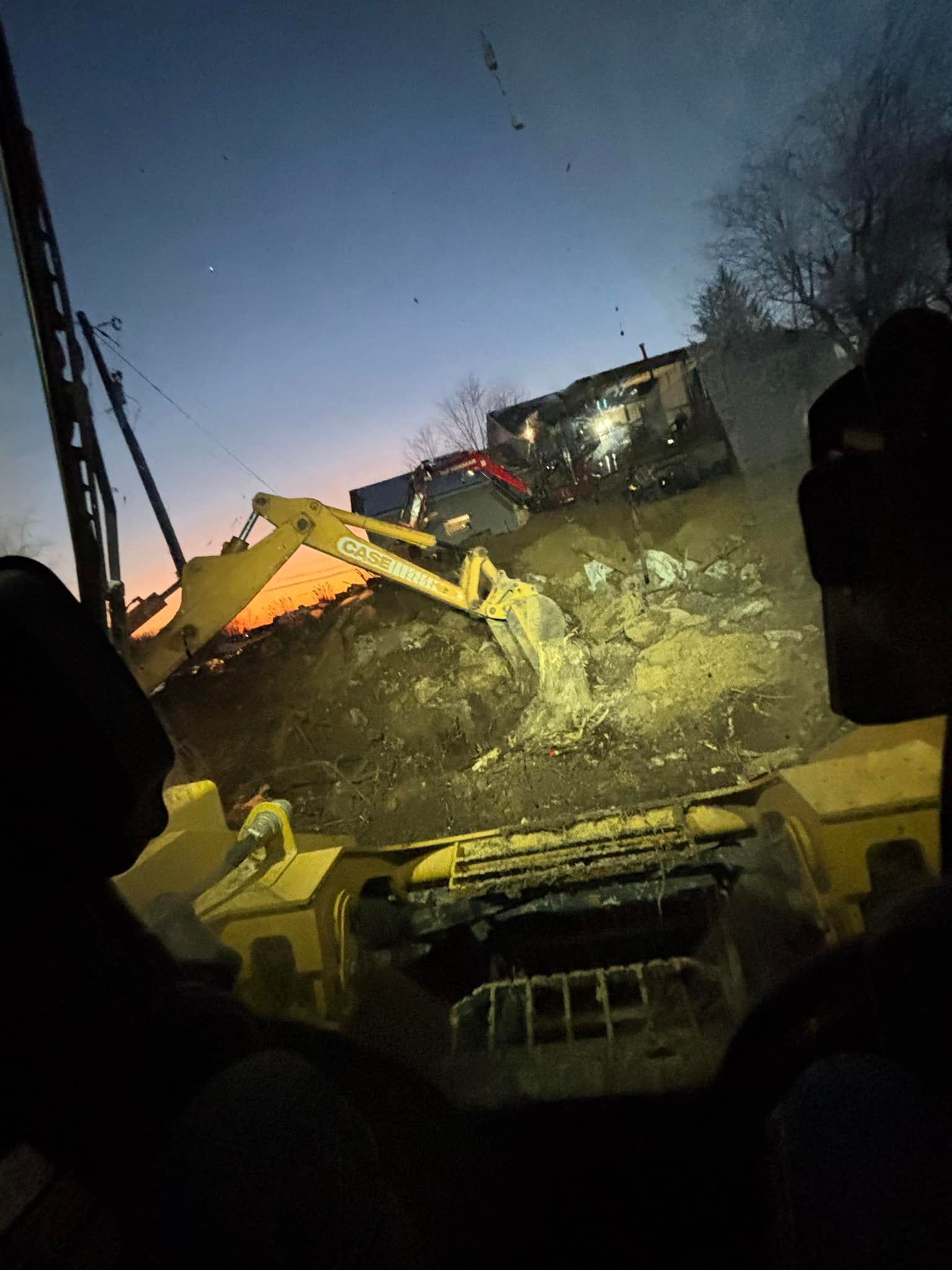 Excavator demolishing a structure at dusk; illuminated by work lights.