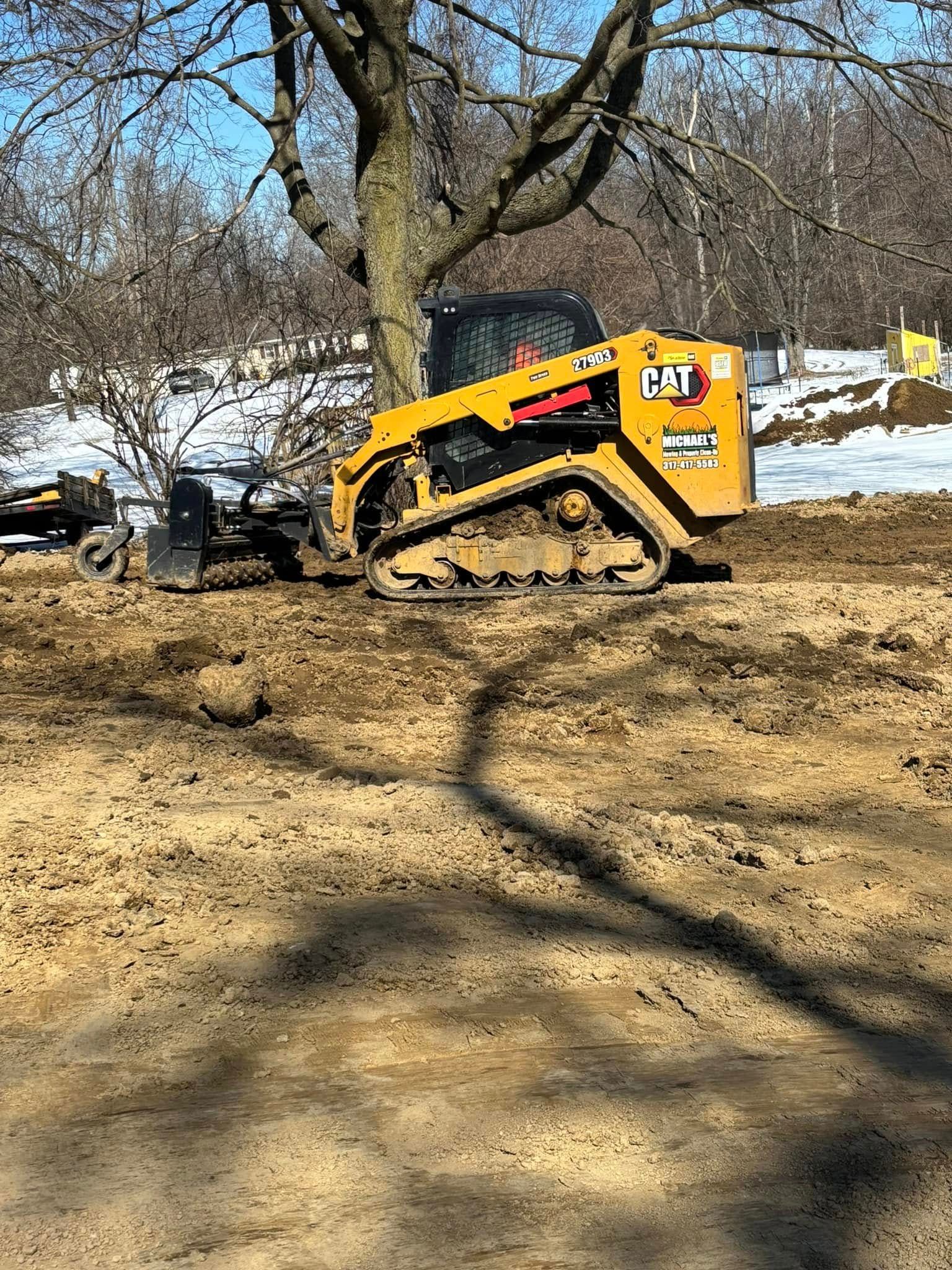 Yellow Caterpillar skid steer on muddy ground near a tree with snow patches in the background.