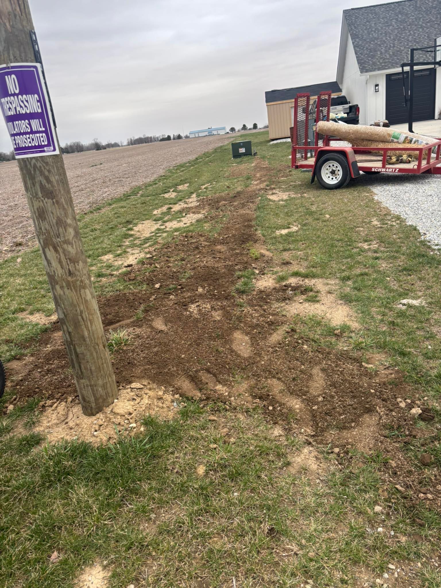 Dirt and damaged grass near a utility pole. A trailer is nearby. A white building and field are in the background.
