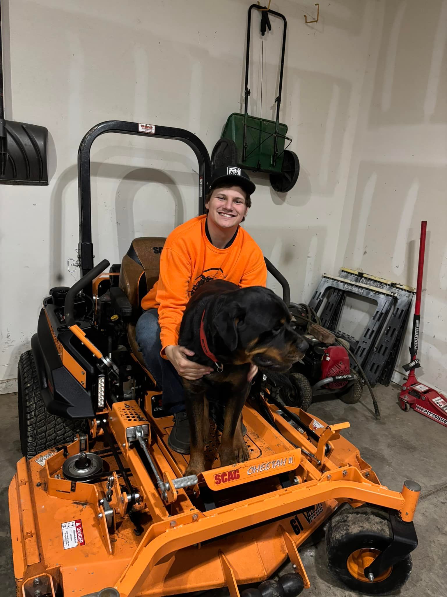 Man in orange shirt with Rottweiler dog on orange lawn mower inside garage.
