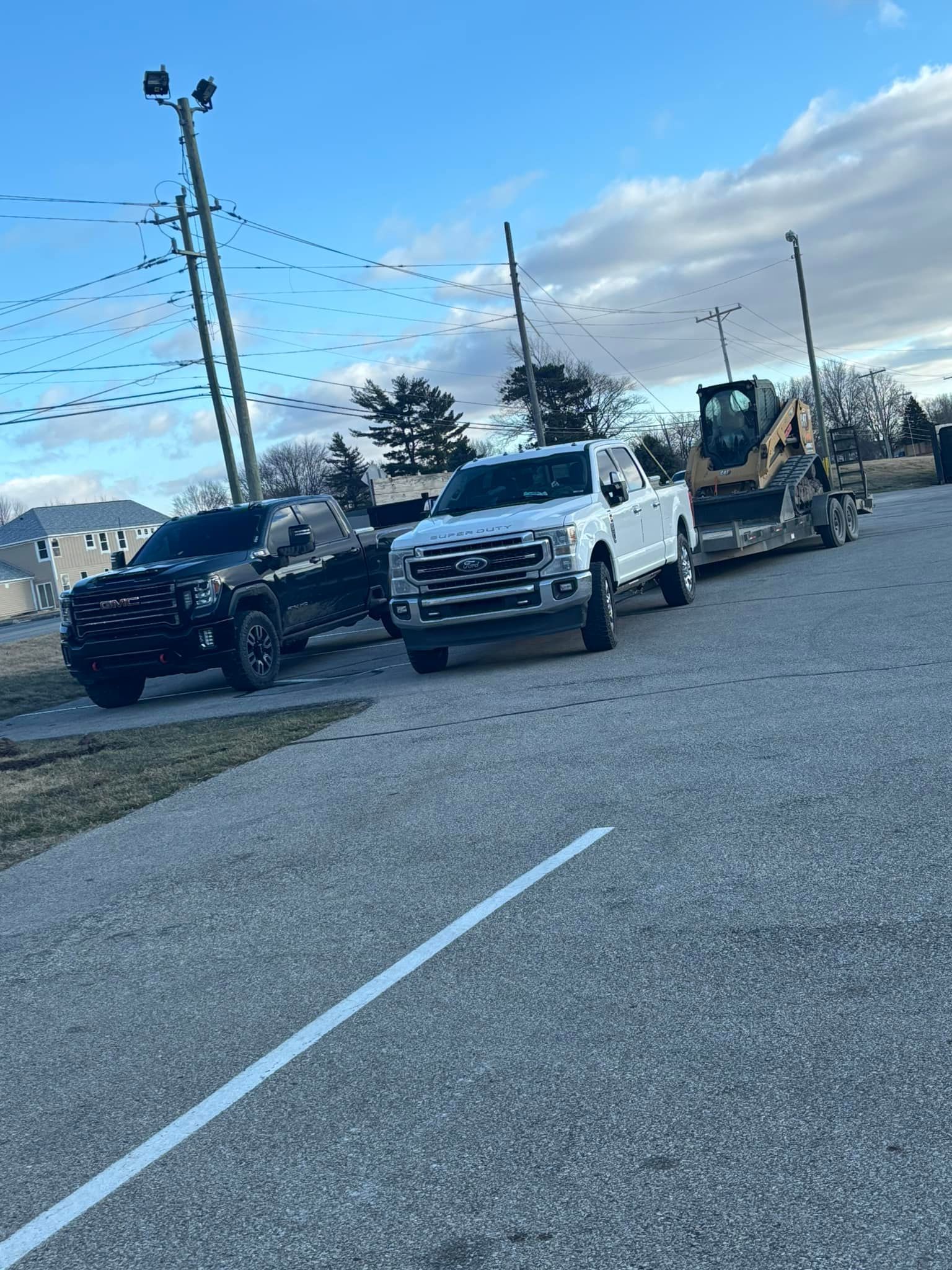 White truck towing a trailer with a small excavator, parked on a paved lot. Black truck is parked to the left.