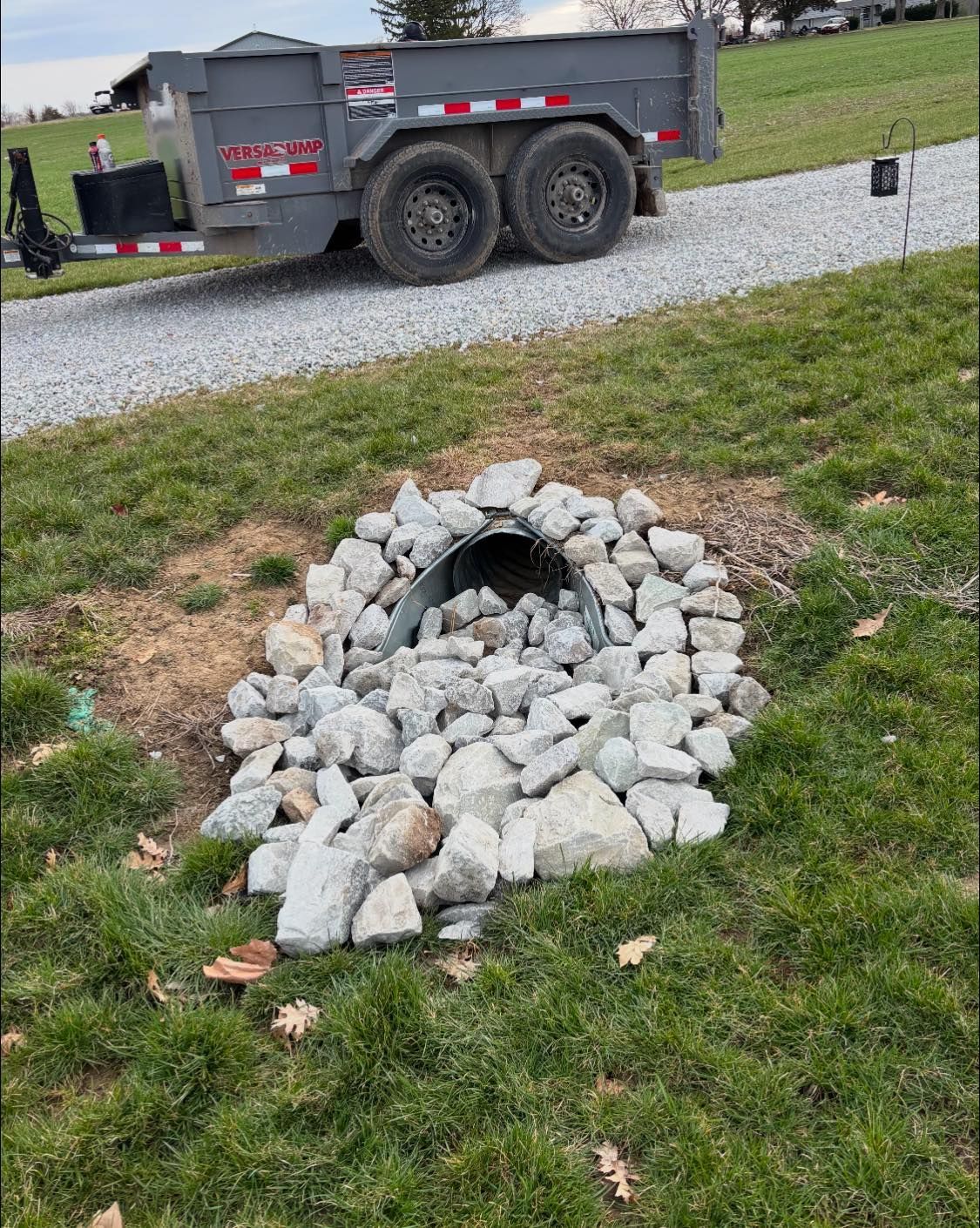 Culvert with rock surround on a grassy slope, with a trailer in the background on a gravel driveway.