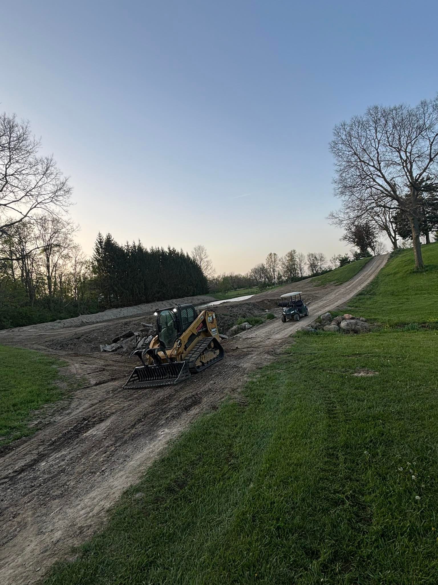 Construction site with small excavator on a dirt road, trees, and grassy areas under a blue sky.