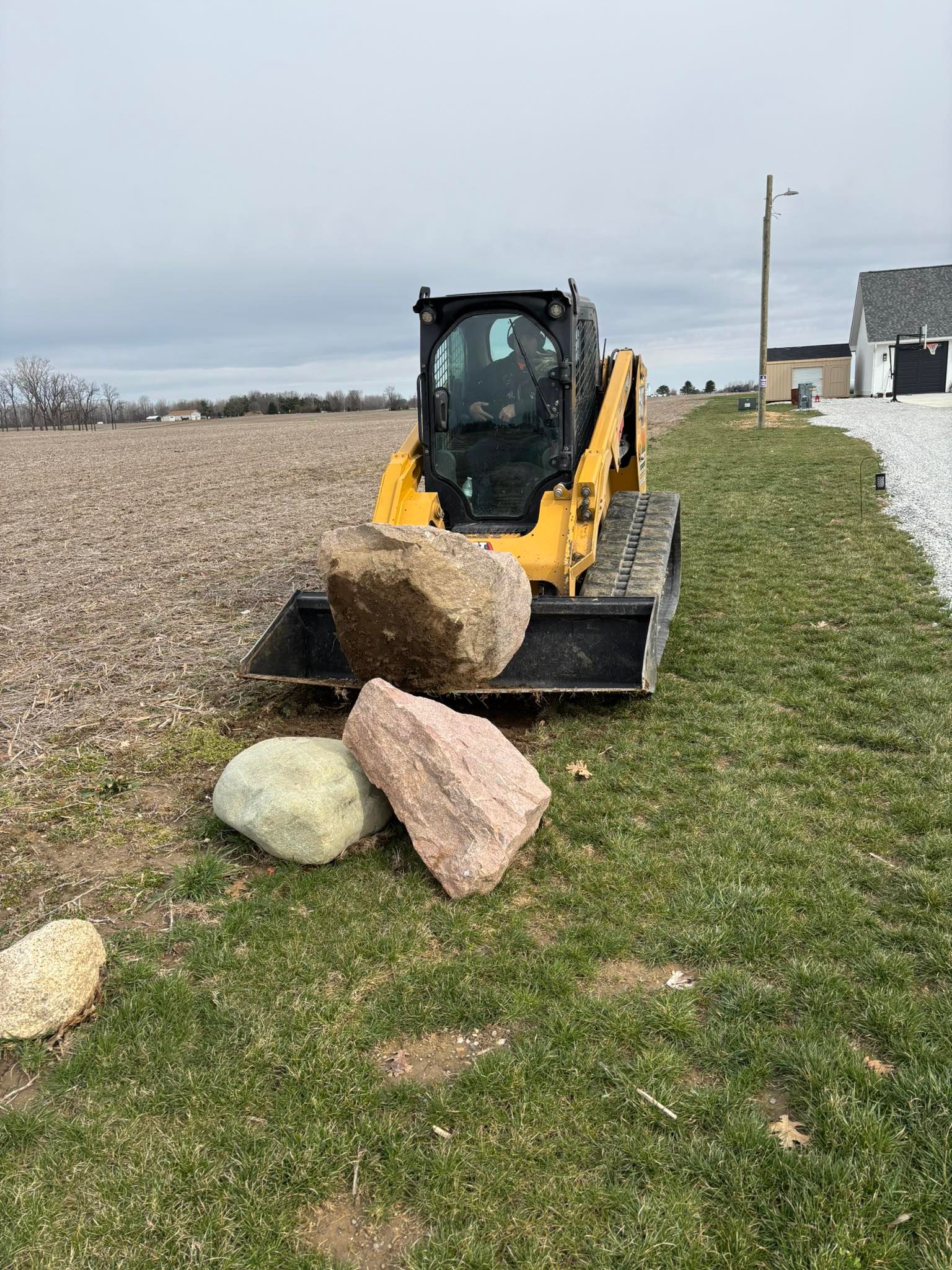 Yellow skid steer loader lifting a large rock near other rocks in a grassy field with a building and sky in the background.