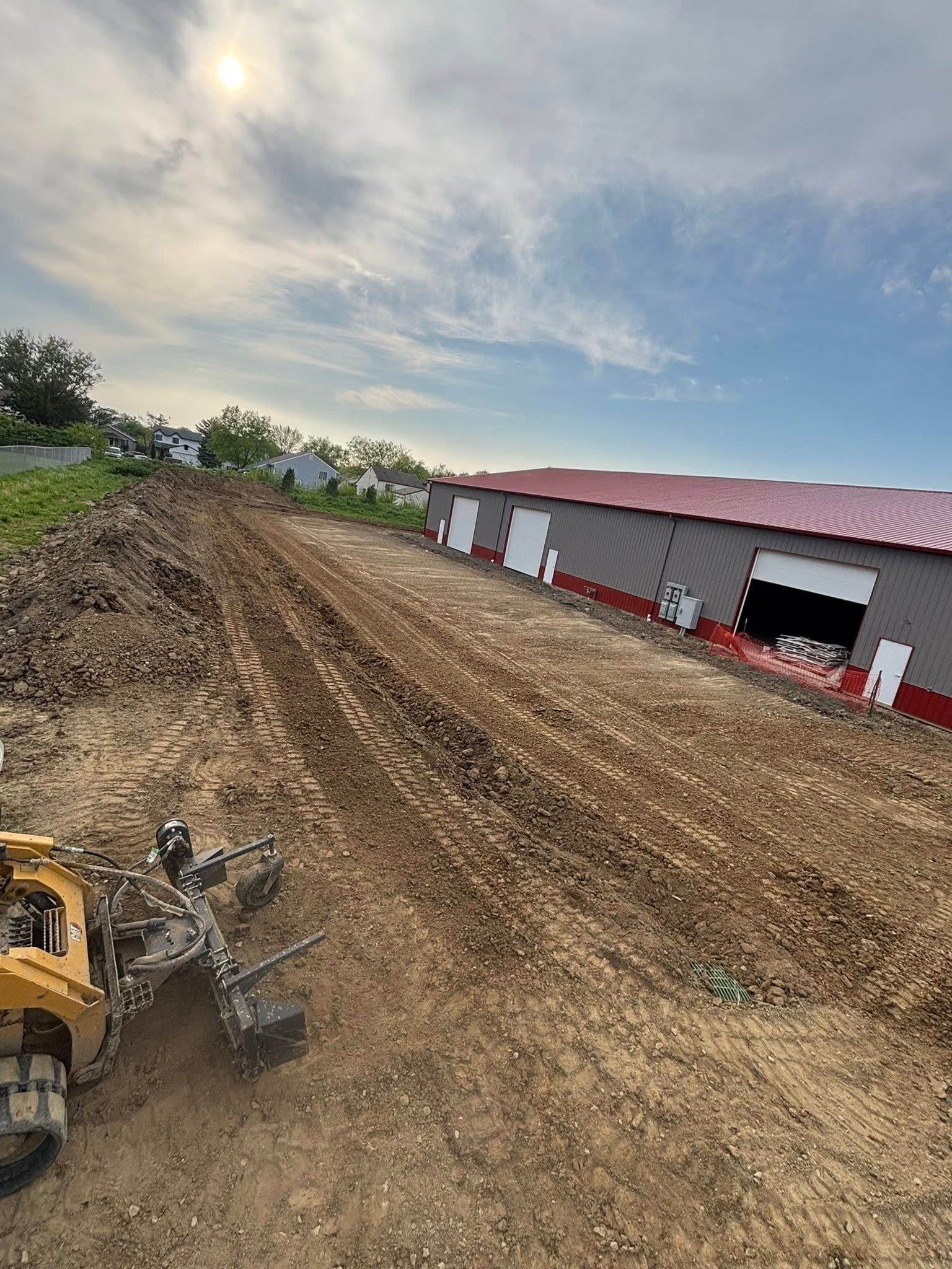 Construction site with a grader preparing a dirt lot next to a commercial building with loading doors.