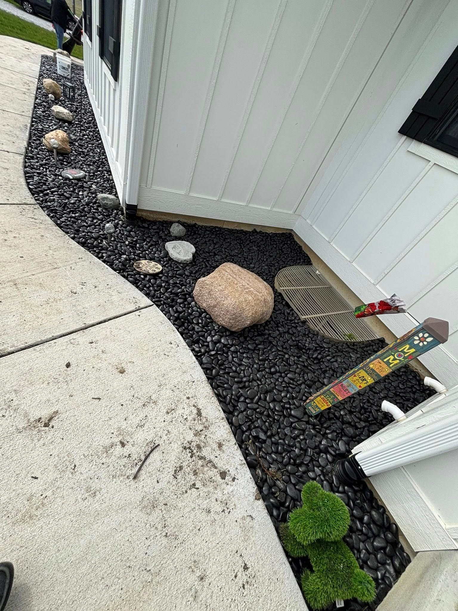 Black rock landscaping beside a white house and sidewalk. Some rocks and a green bush present.