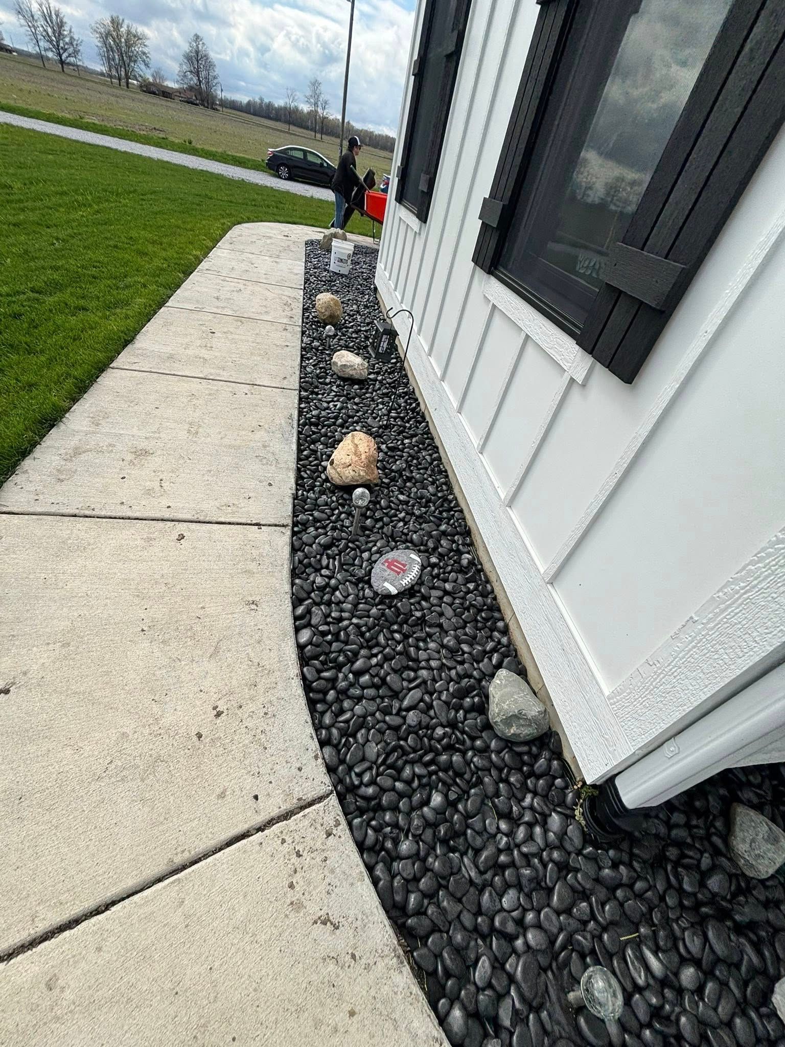 Black rock border along a white building and sidewalk with a few larger rocks interspersed.