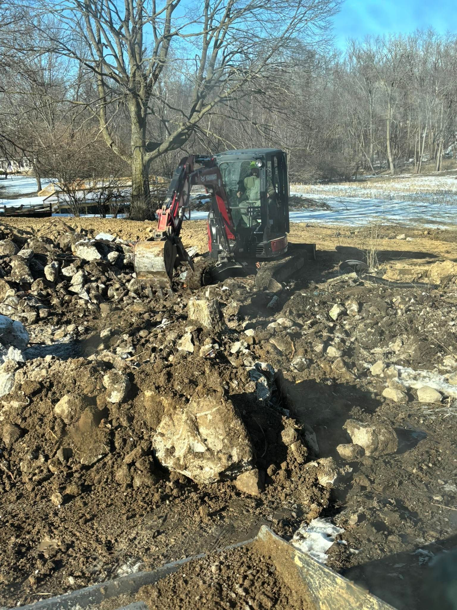 An excavator digging in a muddy, rocky area near a tree and a snowy field.
