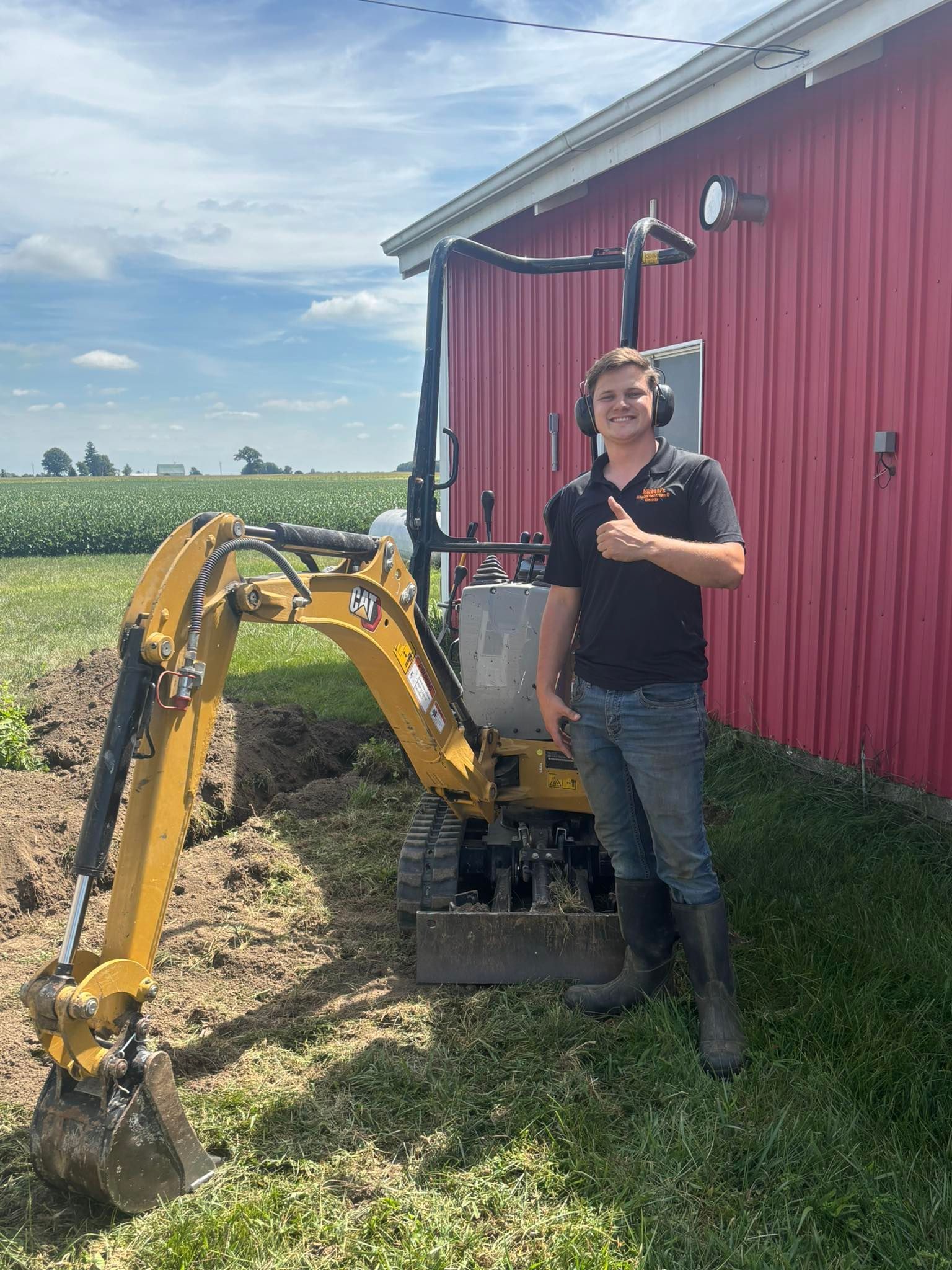 Man giving thumbs up next to yellow excavator, trench next to a red building and field on a sunny day.
