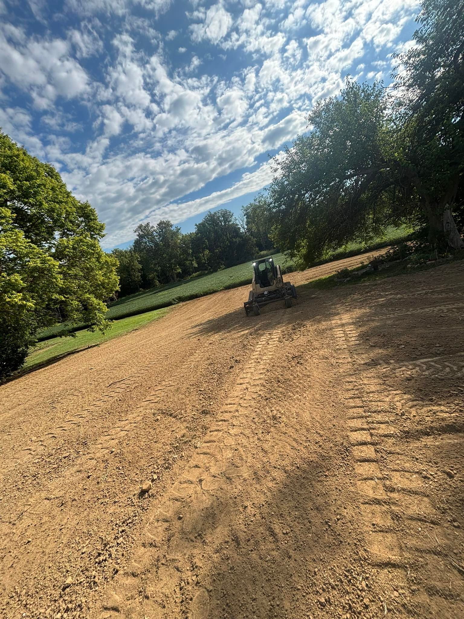 A large graded dirt area with tractor tracks, trees, and cloudy blue sky in the background.
