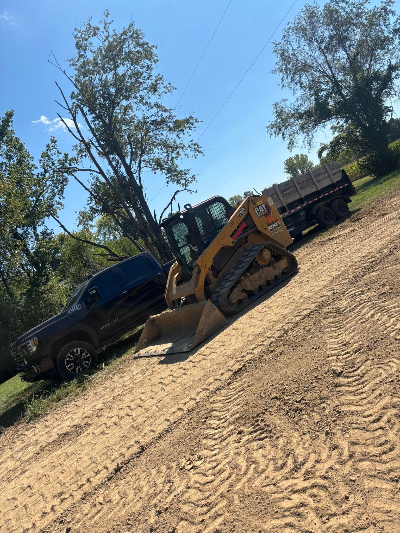 Skid steer loader with a truck and trailer on a dirt road, trees, and blue sky.