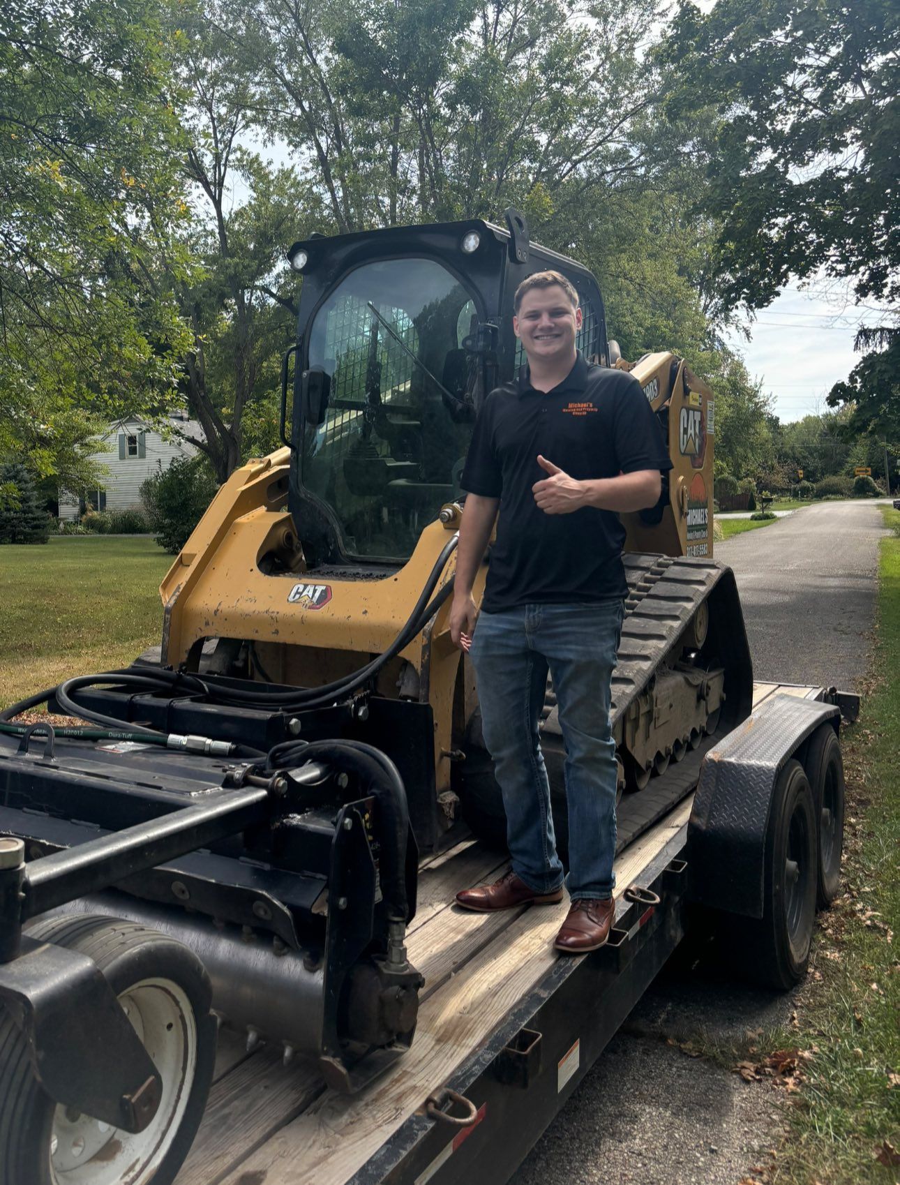 Man standing on a trailer with a yellow skid steer, thumbs up. He's wearing a black shirt and jeans. Outdoors, daytime.