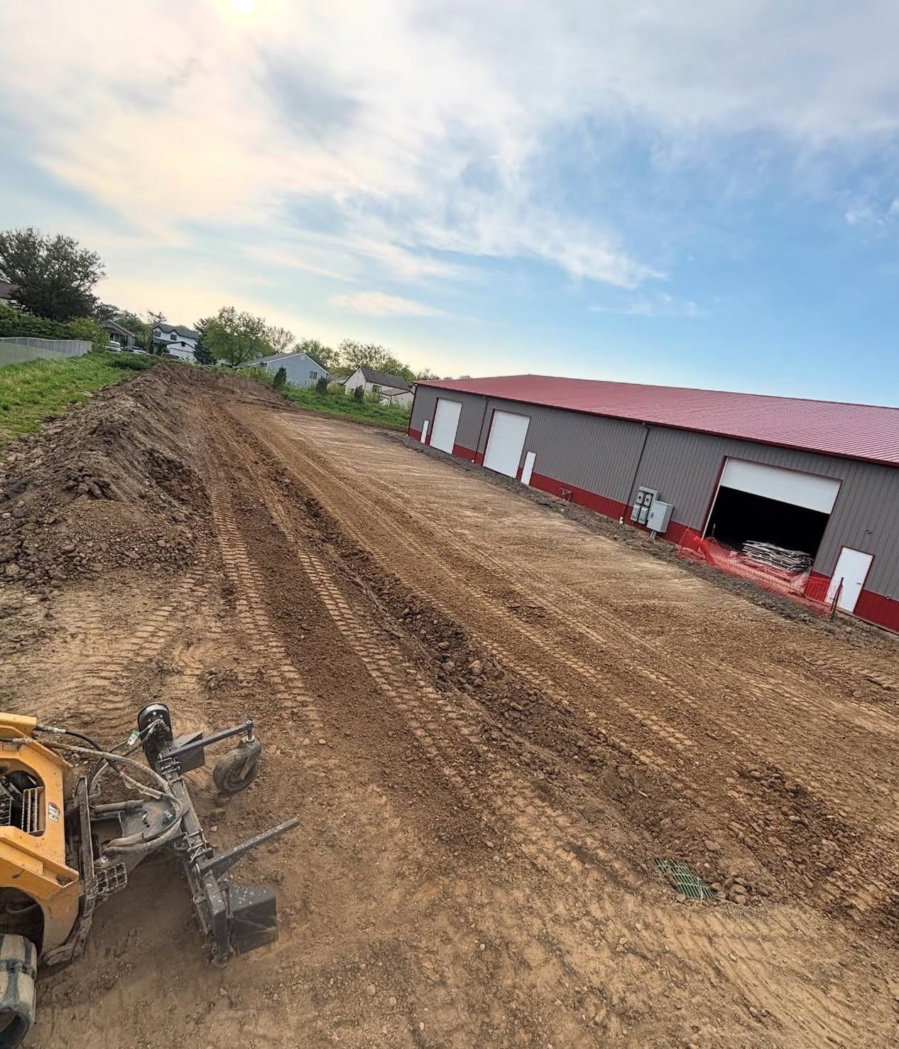 Dirt and construction equipment at a building site with a long dirt path beside a warehouse with white doors.