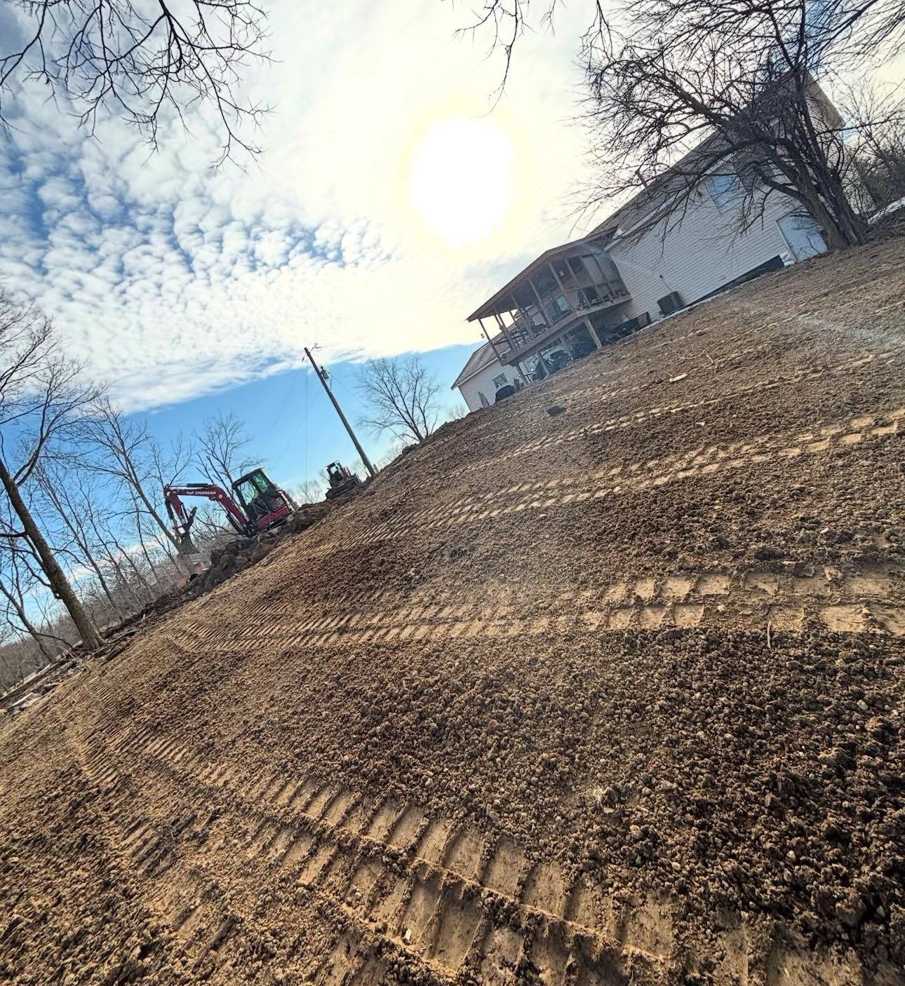 Construction site: Excavator on muddy slope under cloudy sky, house in the background.