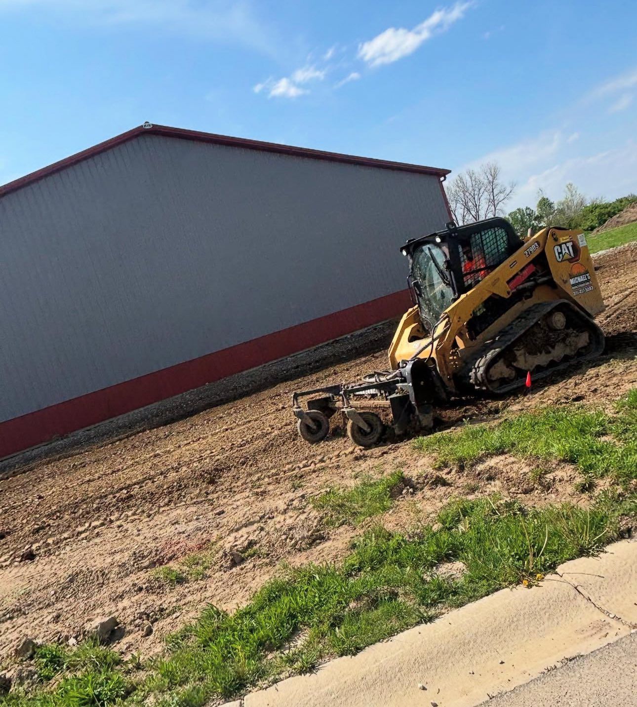 Yellow Caterpillar skid steer grading soil near a gray building on a sunny day.