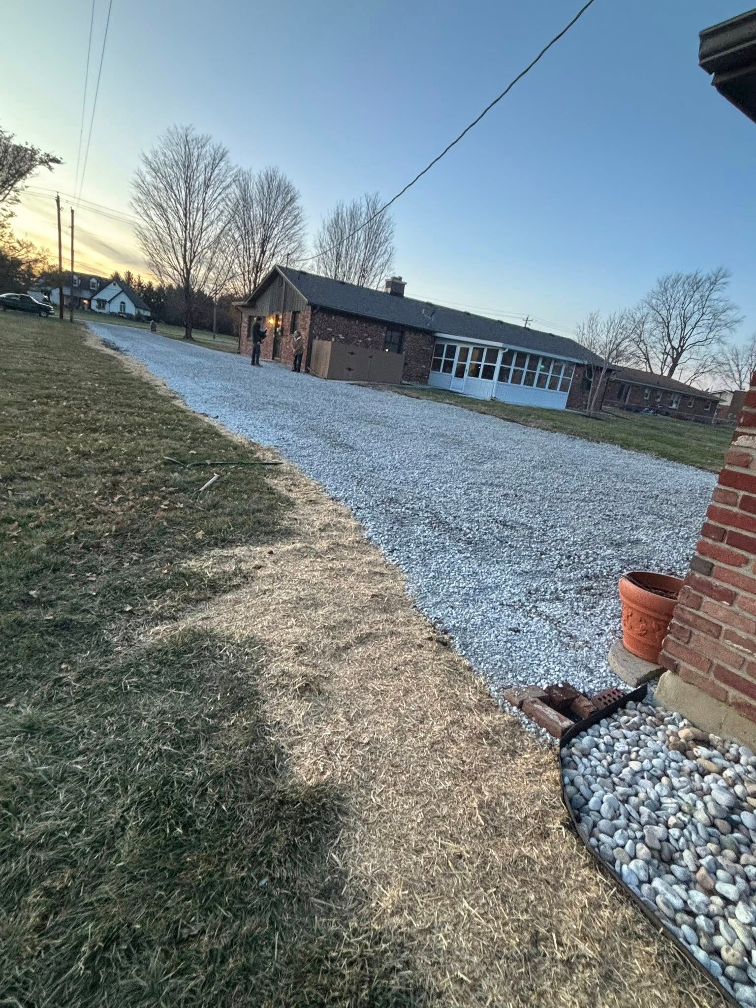 Gravel driveway next to grass and brick wall, leading to a long building and trees under a dusk sky.