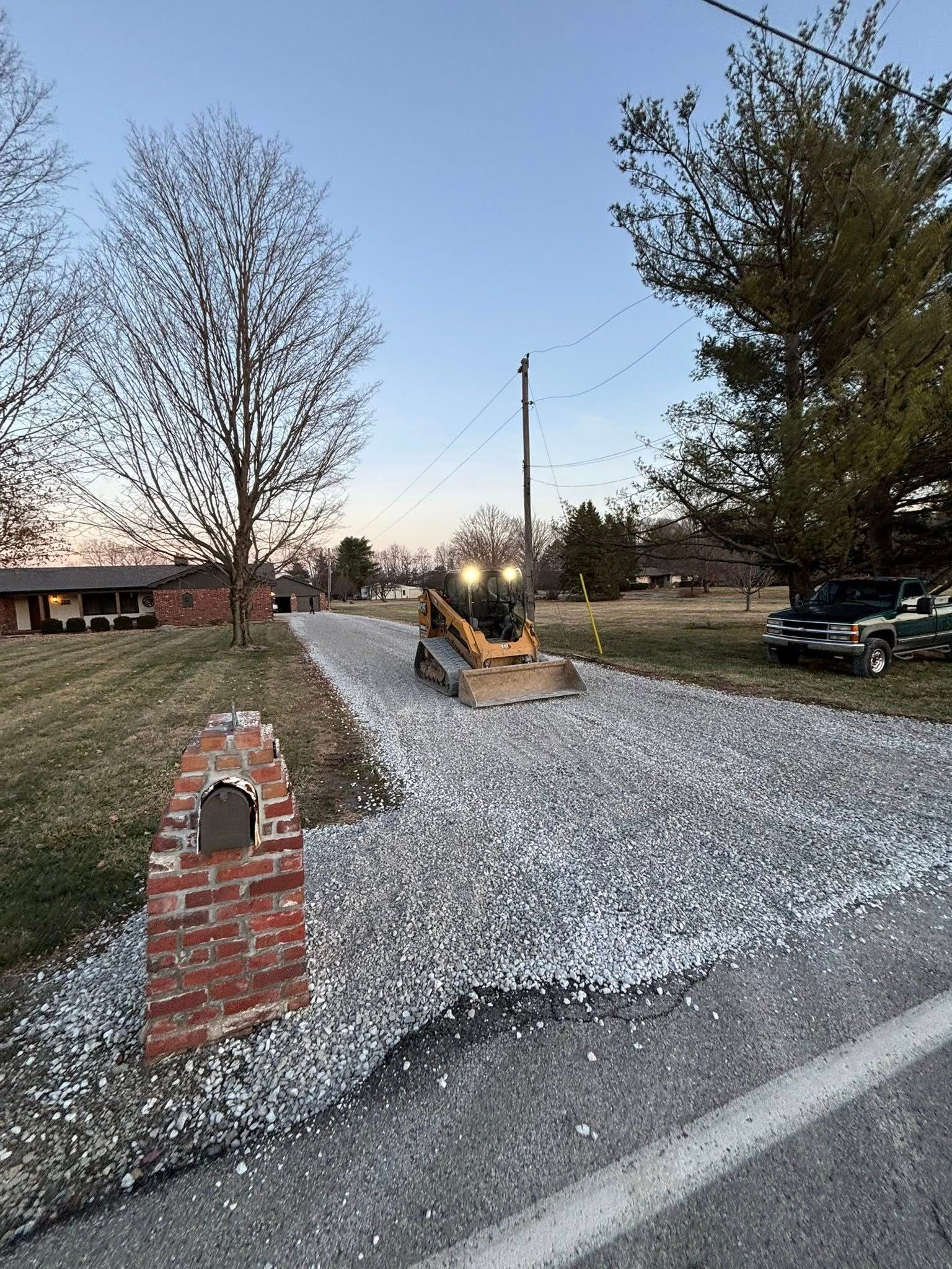 A small construction vehicle leveling gravel on a driveway next to a brick pillar and road, at dusk.