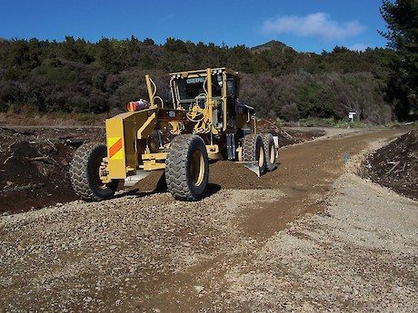 Yellow road grader smoothing gravel on a new road, trees in the background.