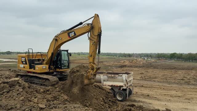 Yellow excavator loading dirt into a white trailer on a construction site. Cloudy sky in the background.