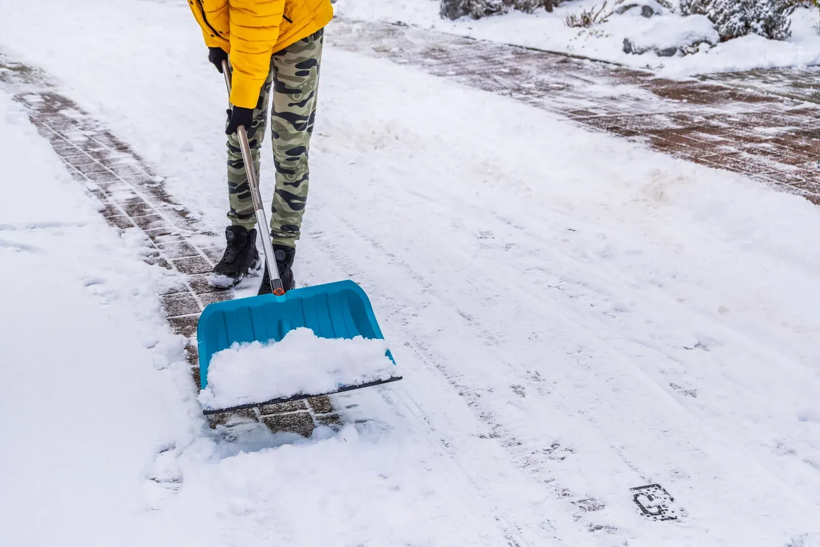Person shoveling snow on a snowy driveway, wearing a yellow jacket and camo pants.