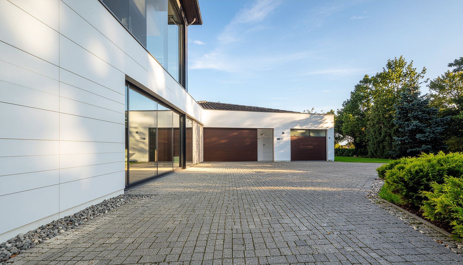 Modern white building with dark wood garage doors, gray brick driveway, and landscaping.