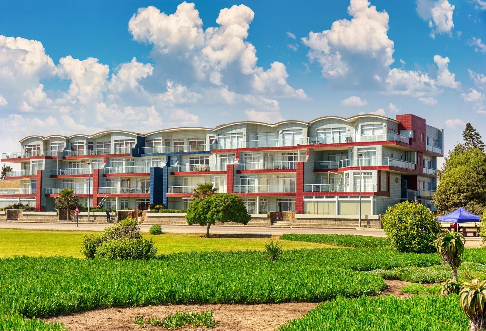 Multi-story building with balconies, blue and red accents, against a sunny sky, lawn in the foreground.