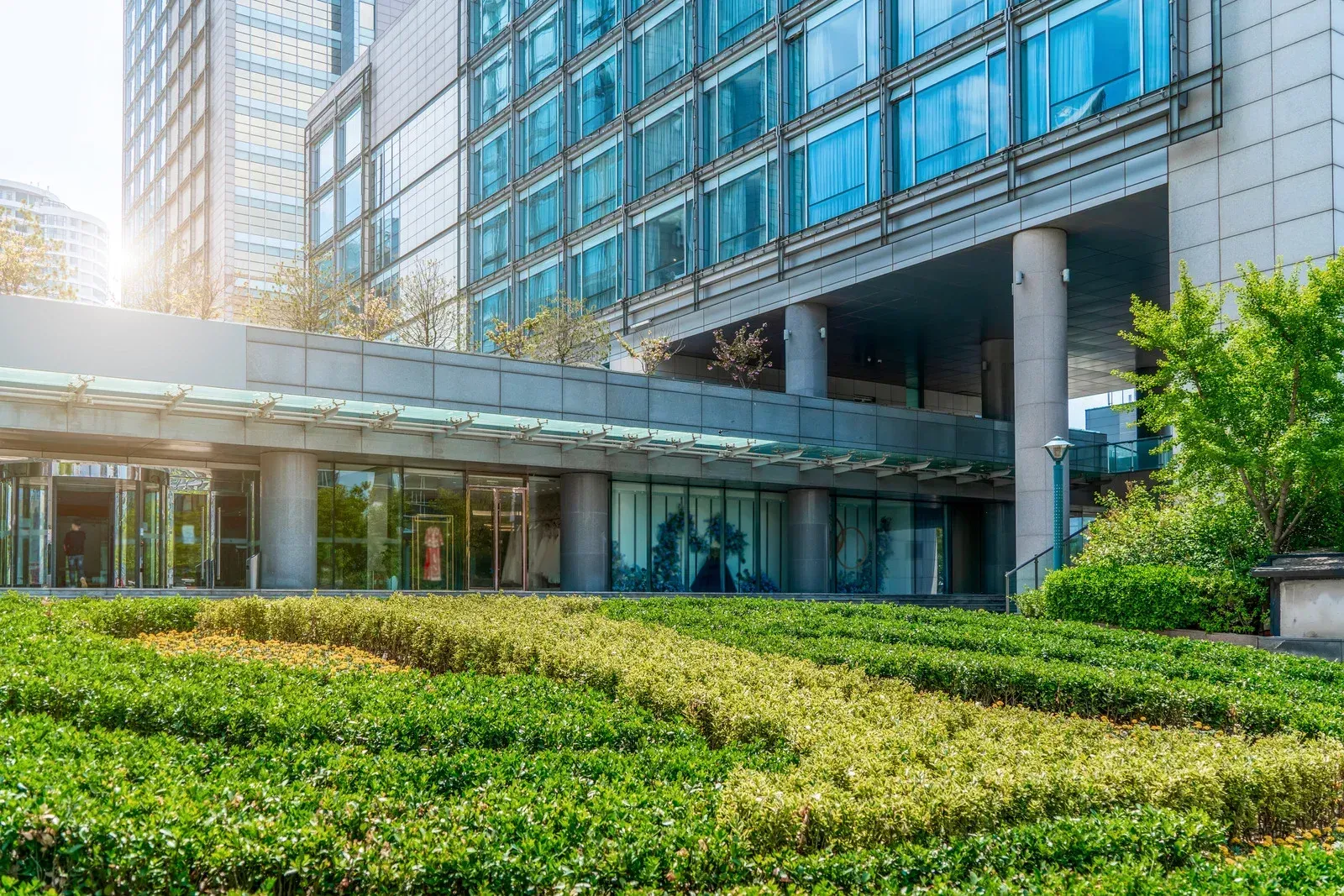 Modern building entrance with glass doors, lush green landscaping, and blue-tinted windows.
