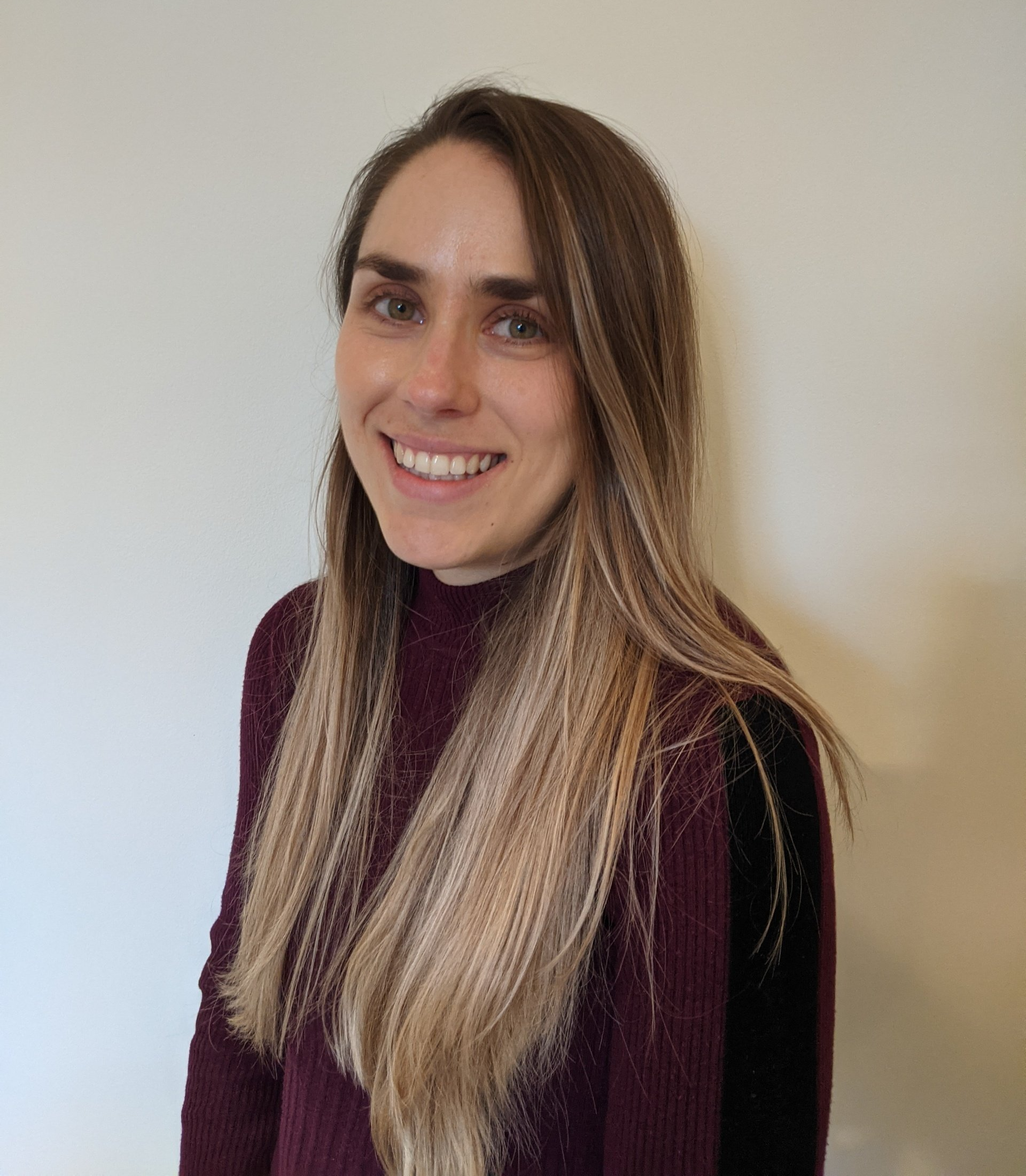 A woman with long blonde hair is smiling and wearing a purple sweater.