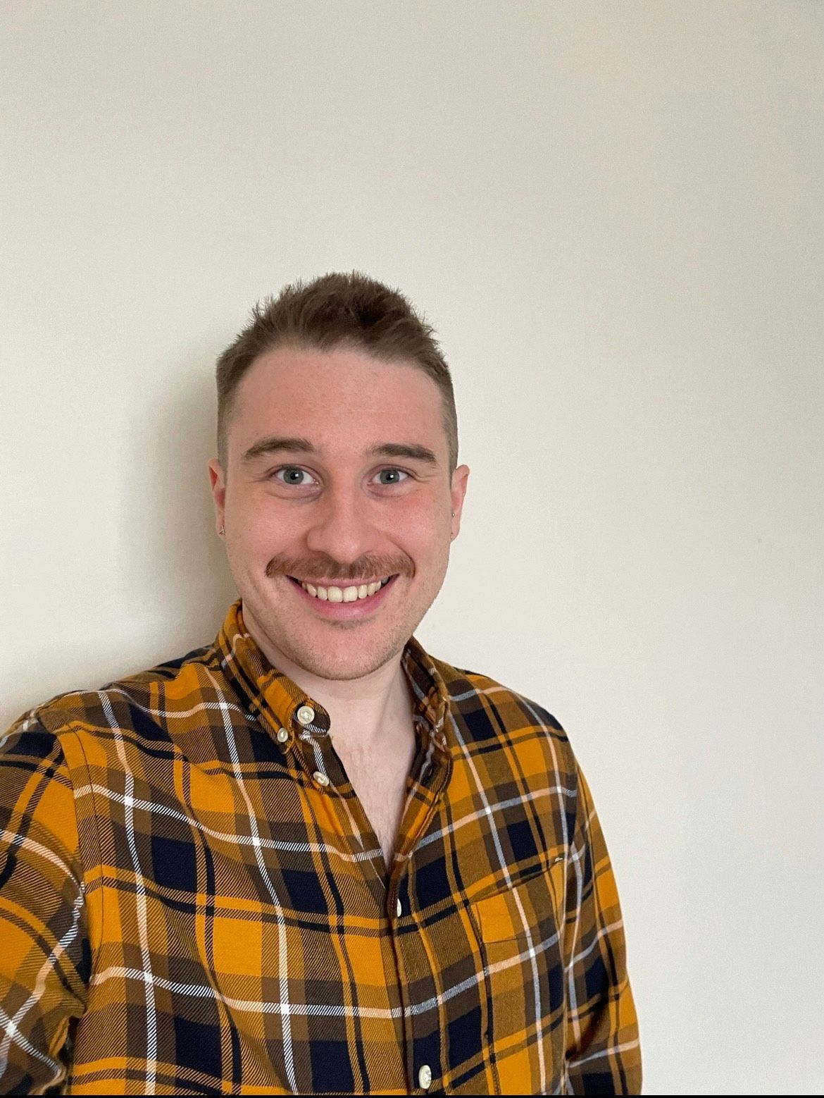 A man in a plaid shirt is smiling and standing in front of a white wall.