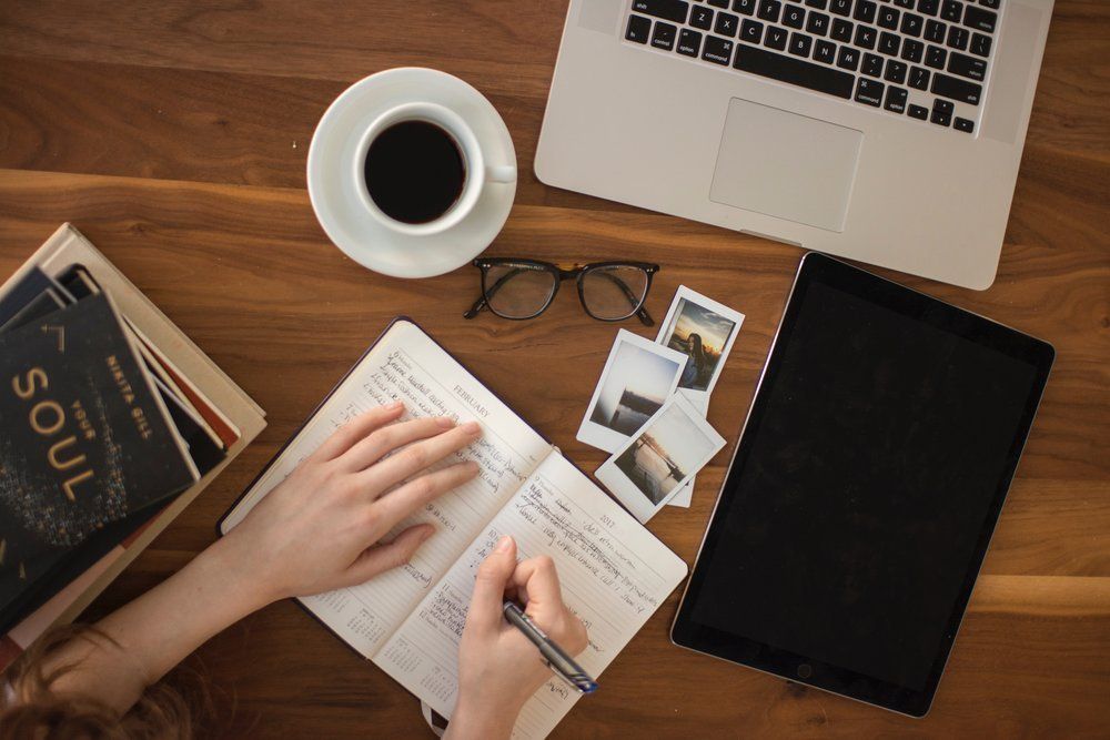 A person is writing in a notebook next to a laptop and a tablet.