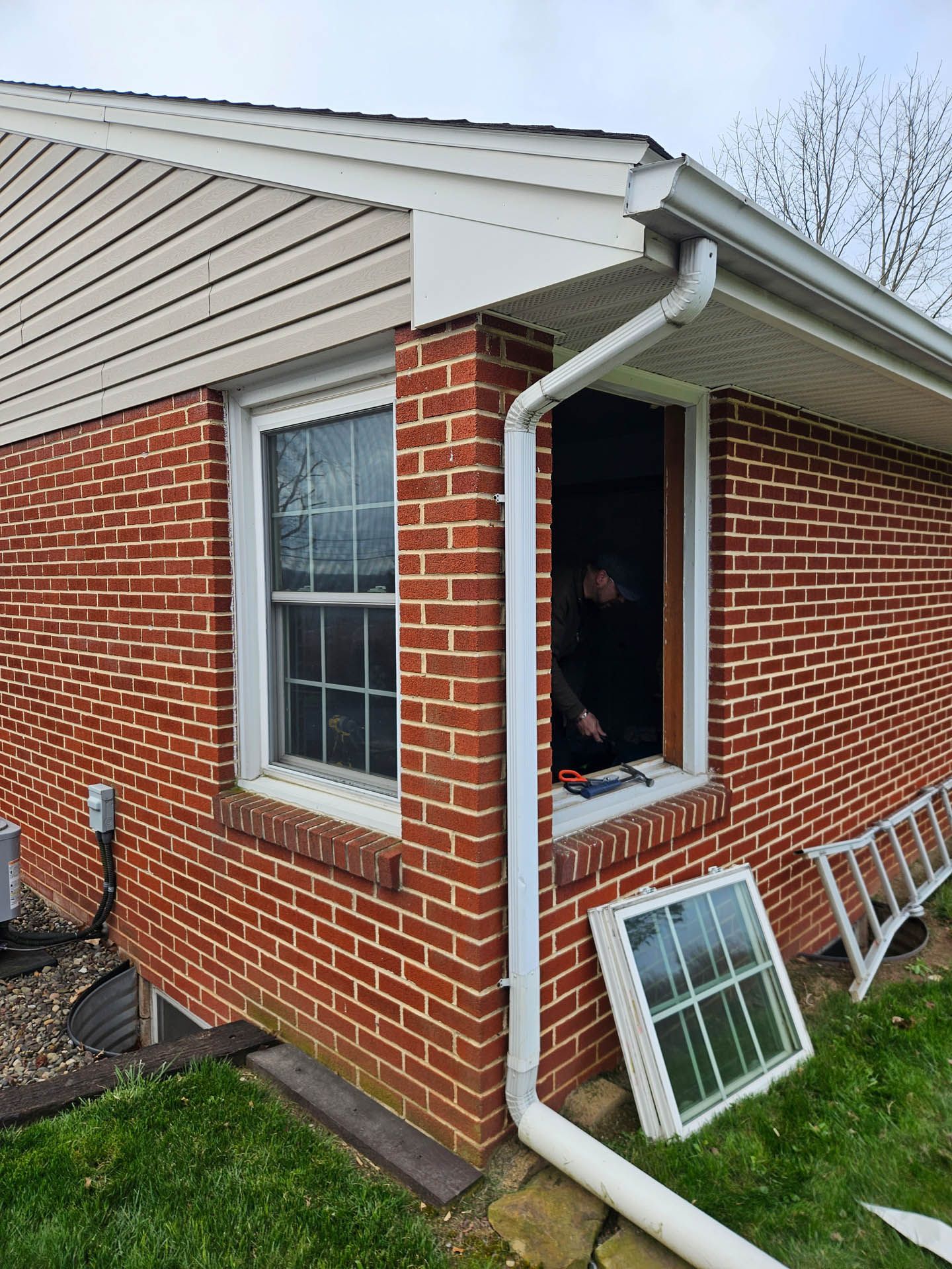 Brick house corner with open window and a white gutter; window screens lean against the wall on the grass.