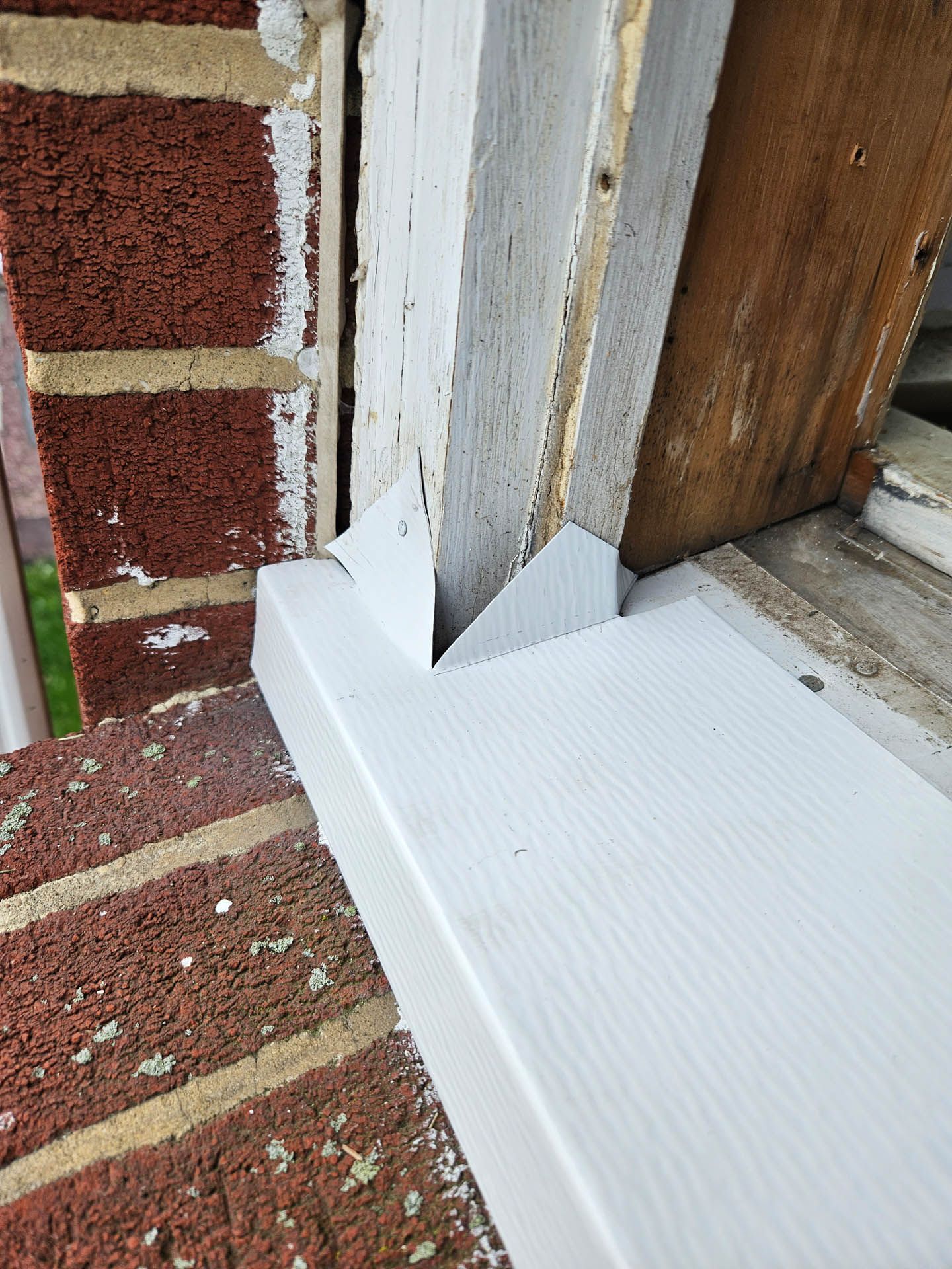 White flashing installed around a wooden door frame beside a red brick wall and window sill.