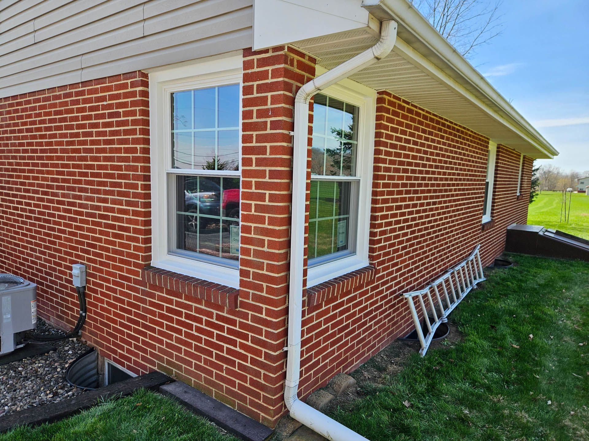 Brick house corner with white-trimmed windows and gutters, lawn beside it, and a ladder leaning against the wall