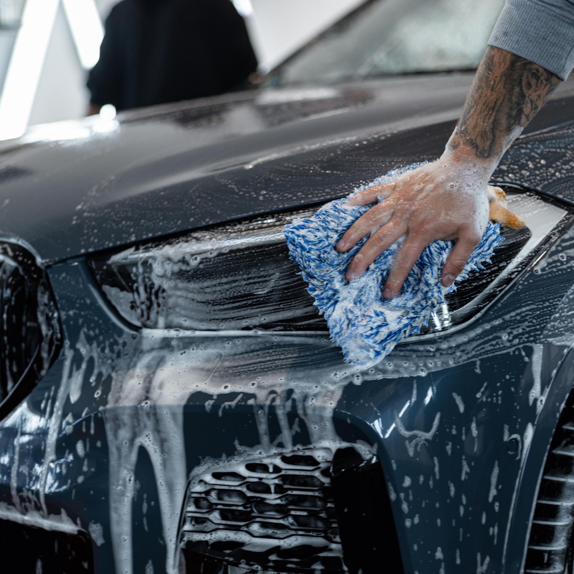 Person washing a dark-colored car with a blue cloth and soapy water, close-up on the front.