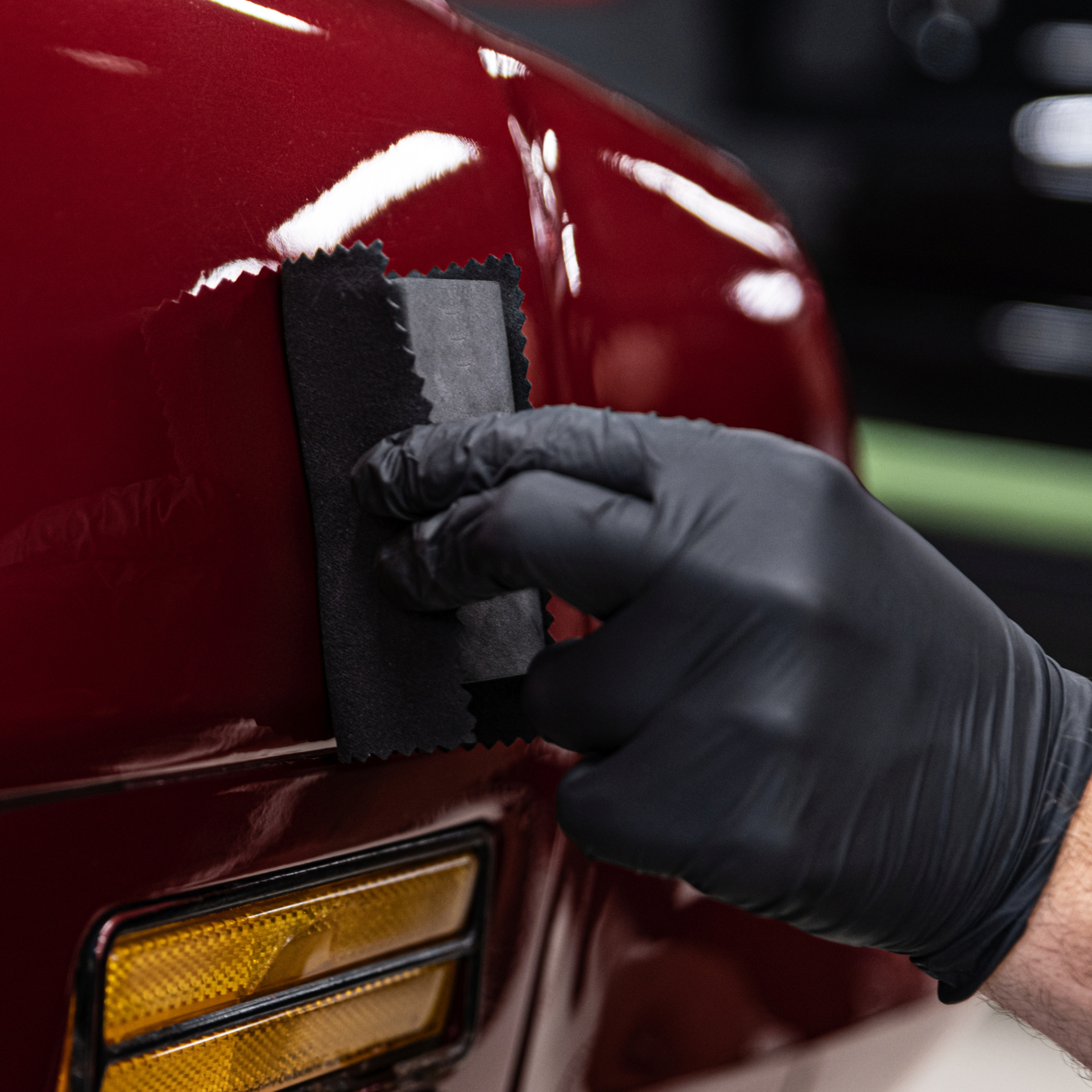 Gloved hand applying a coating to a red car panel with an applicator.