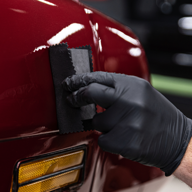 Gloved hand applying a coating to a red car panel with an applicator.